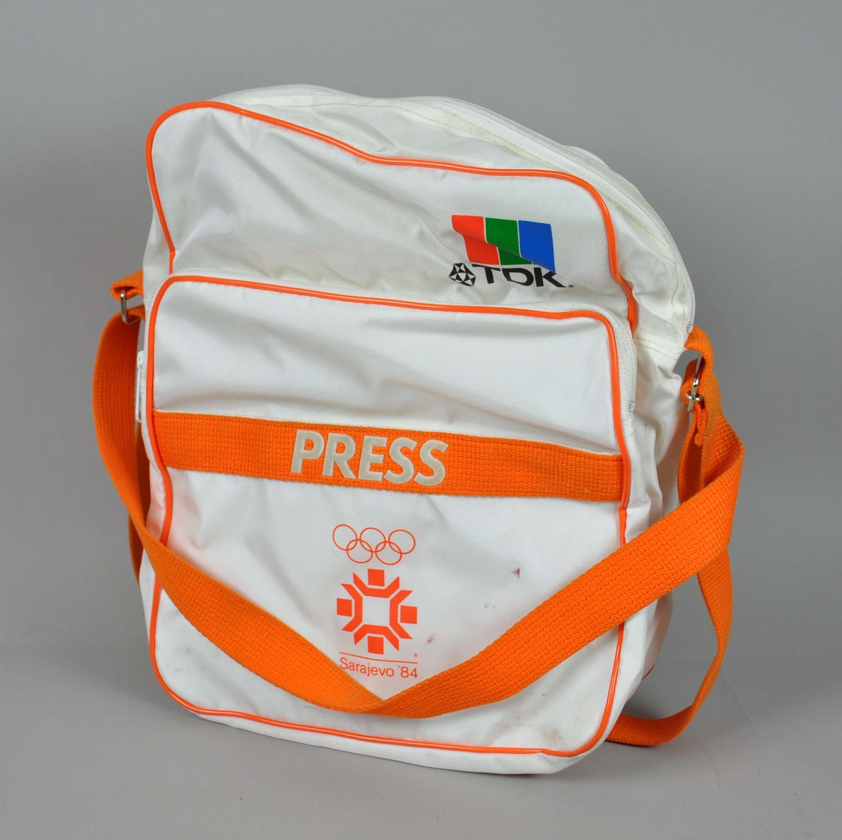 Hvit og oransje bag med skulderreim. På bagen er det emblem for de olympiske vinterleker i Sarajevo i 1984 og en logo for TDK.