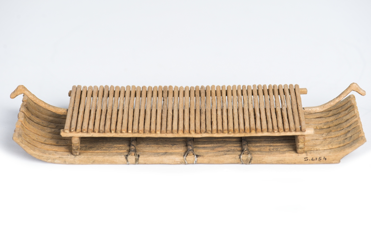 Modell av flotte, rekonstruktion av "Bredarörtypen", skeppsbild från Bredarör invid Kivik, Skåne. Flytkropp av 9 raka, i ändarna svagt uppböjda stockar, sammanhållna av 3 tvärbalkar och två plattformsstöttor. Plattformen består av tvärgående ribbor uppburna av tre längsgående spärrar varav den mittersta, utskjutande, avslutas med djurhuvud. Skala: 1/60 (Jfr. S.5892 = skala 1/50).