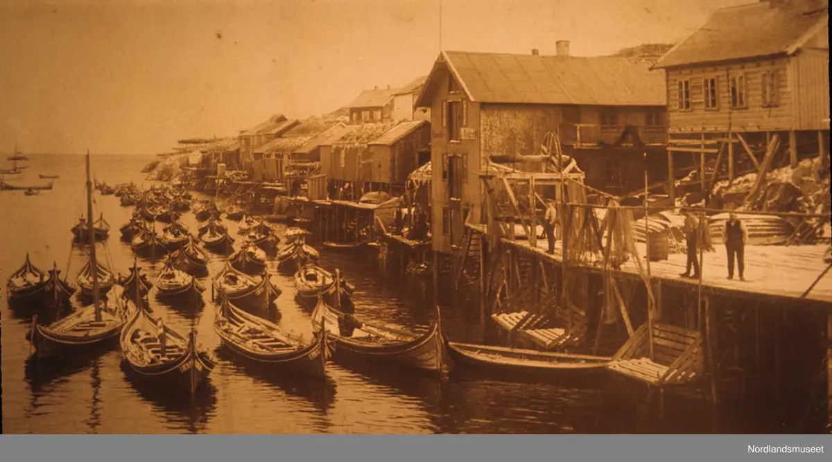 Ballstad, kaianlegg med tilhørende bygninger t.h. Flere personer på kaia. På havna ligger flere nordlandsbåter av forskjellig størrelse.