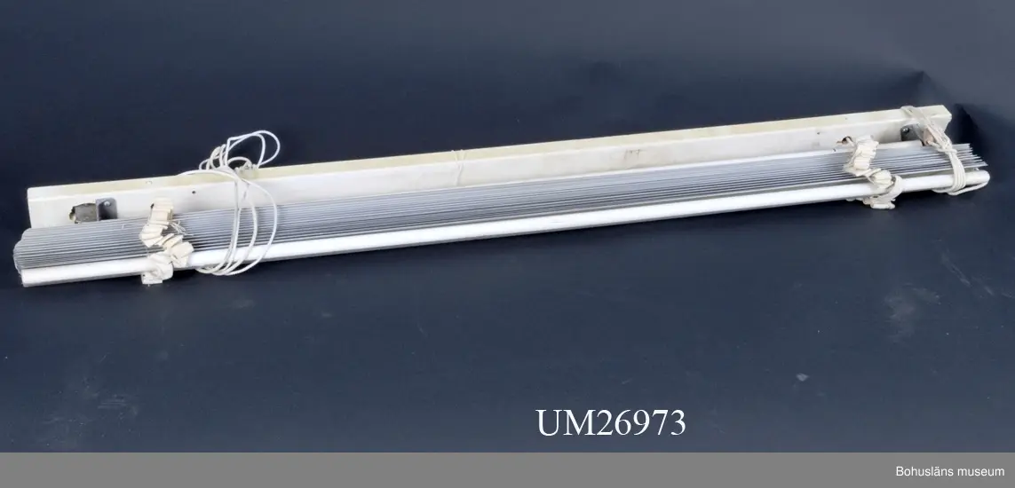 471 Tillverkningstid 1930-60
410 Mått/Vikt !B 114 L UPPDRAGEN 15 CM *
410 TJ 5 CM
601 Gråtonat vit persienn. Bräda av trä med metallbeslag där översta
603 lamellen, av trä, är fastsatt. På ena sidan finns en trissa där snöre
6031 av bomull kan löpa. Nedre lamellen är också av trä men övriga av
6032 metall. Lamellerna är fästa vid varandra med bomullsband och snöre
6033 löper på vardera sidan genom hål i lamellerna. På sidan utan trissa
6034 upptill är det snöre av syntetfiber.
6035 På ena sidan finns två träkläppar fastknutna på snörändar.
6036 Något smutsig.
6037 Inlämnad till museet av Eskil Fredriksson som arbetar på KVC (Kultur-
6038 vårdscentrum). Den har suttit i en fastighet i Ljungskile och är läm-
6039 nad till Eskil av hans svåger som skruvat ner den då hans dotter och
60391 mor köpte fastigheten, eftersom svågern ansåg att det var synd att
60392 slänga en såpass gammal persienn. Eskil själv hade varit på skol?resa
60393 till Värmland år 1948 och sett en utställning om persienntillverkning.
60394 Han mindes att på utställningen presenterades persienner som en nyhet.
60395 I tidskriften "Hem i Sverige Tidskrift för Bygge Trädgård och Hem-
604 kultur" (se museets bibliotek)  finns "Hem i Sveriges inköpstjänst"
605 dvs sidor med adresser till olika firmor som kan bidra till husbygget.
606 Där tas Metallpersienner upp för första gången 1942.
607 Med all sannolikhet har persienner tillverkats längre i Sverige, men
608 att ordet persienn eller metallpersienn inte förekom i tidigare år-
609 gångars inköpstjänst kan möjligen betyda att föremålet tidigare inte
610 var så känt eller efterfrågat.
6101 I svensk uppslagsbok från 1937 finns ordet persienn som uppslagsord.
6102 Inget står dock om persienntillverkning.
6104 Intresset för boende och heminredning var stort i samhället redan
611 under 1930-talet och med ideer om att husen skulle byggas så att luft
612 och ljus skulle släppas in i lägenheterna kan behovet att avskärma sig
613 mot solen också ha kommit att öka. (Det senare är funderingar vid
614 vid katalogiseringen). Uppgifterna från Eskil Fredriksson är från ett
615 telefonsamtal 1999-08-18.