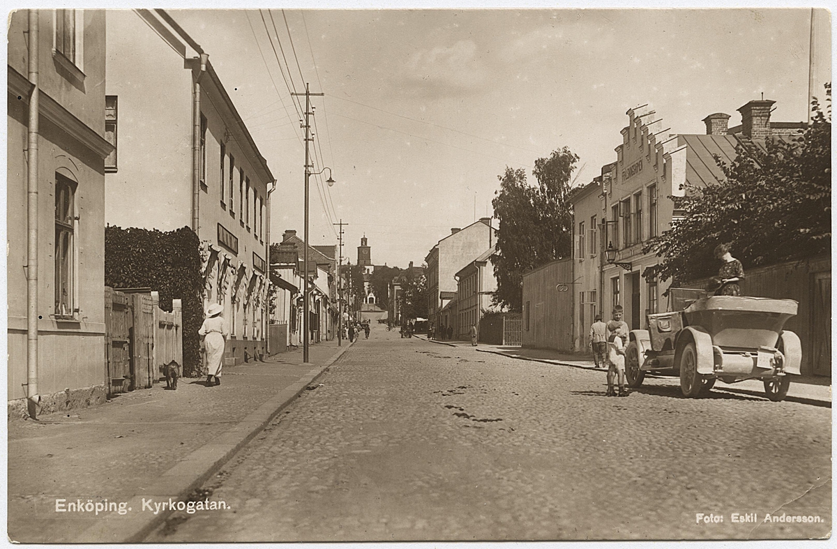 Kyrkogatan sett från sydväst, Enköping, mitten eller senare hälften av 1920-talet.
Påskrift på baksidan: "1925? Mamma Hedvig stående i bilen, en Scania-Vabis med växelspakarna utanför. På gatan vid bilen bröderna Bror o Nils Thulin."