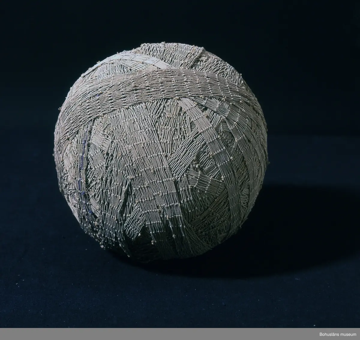 Runt, grå-beige garnnystan bestående av kasserat fisknät.
När gamla fisknät var uttjänta återanvändes de ofta till mattor.
Att klippa mattrasor av garn var en sysselsättning för hela familjen - gamla, unga, kvinnor och män.
Mattor med inslag av nät var starka och låg bra på golvet enligt givaren. Mattorna hade inslag av både nät och vanliga trasor. Det var viktigt att remsorna klipptes jämt. En del av nätet knöts fast i kökssoffan för att hålla det sträckt vilket gjorde det lättare att räkna stolpar och klippa efter dem och på så sätt få jämnbreda remsor. Nätet klipptes längs med den sida som inte var fäst i soffan och allt eftersom arbetet fortgick flyttades nätets fästpunkt.
På en "vad" var ca 15 - 20 stolpar en vanlig bredd. Vaden hade mindre maskor än nätet vilket innebar att det blev ett tätare inslag. "Vad" var ofta färgat i blått eller grått och garnen ofta i olika bruna nyanser.
Mattorna gjordes rent i saltvatten på så sätt blev de styva och låg plant på golvet och saltet förhindrade även färgerna att färga av sig.

Brukaren är givarens mor. Eventuellt kan nystanet vara tänkt att användas av givarens farmor som också bodde på Smögen.
Tillverkaren är trolig. Han var givarens bror och mycket duktig på att klippa mattrasor av fisknät.

Maria Rödström, gift med Karl-Olof Rödström, f. 1916.
Karl-Olof Rödström var son till Evert Rödström, f. 1898 och Irene Rödström (1903 - 1995).
Evert och Irene är också föräldrar till Karin Rödström. 
Evert är bror till Gertrud Linnea Gunnarsson, f. 1896 med dottern Berit Gunnarsson, f. 1922.
Evert och Gertrud Linneas föräldrar är Frans Rödström, f. 1871 och Sofia Rödström , f. 1872 
Se flera föremål från Smögen i samlingarna från denna Smögenfamilj.
Intervju med Maria Rödström förvaras under Intervjuserien, Etnologi, Ämbetsarkivet.