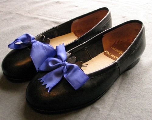 Ett par lågklackade svarta damskor i skinn av typen tysksko, ballerinasko. Blå sidenrosett fastknuten på tåhättan. Taggig, kort "tunga" in mot foten under rosetten. Lädersula och läderklack. Invändig ljusbrun skinnplatta i hälen med guldtryck; "Skoparet Falkenberg".
Gjorda efter original på Länsmuseet i Kristianstad.