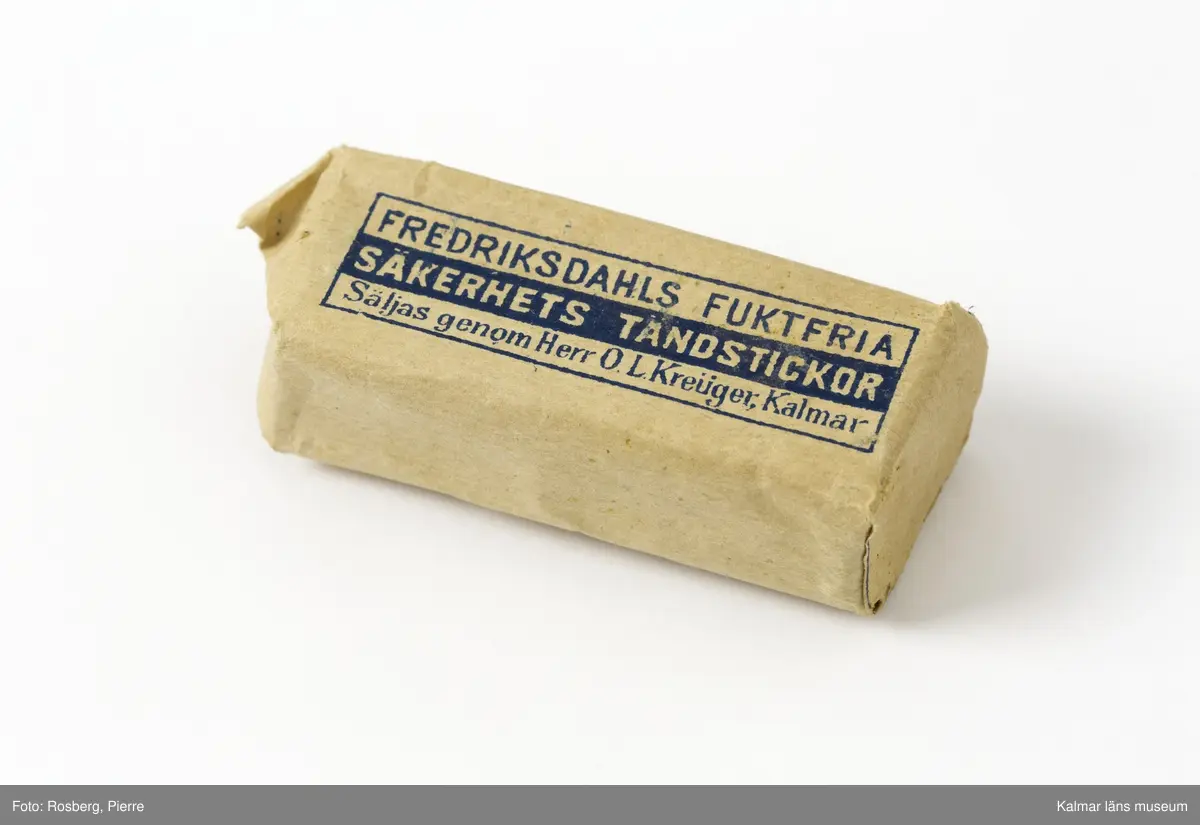 KLM 45567. Tändsticksask, av papper. Tändstickor inslagna i ett emballage av brunt papper, med text i blått: Fredriksdahls Fuktfria Säkerhets Tändstickor, Säljs genom Herr O L Kreüger, Kalmar. På undersidan ett tändsticksplån.