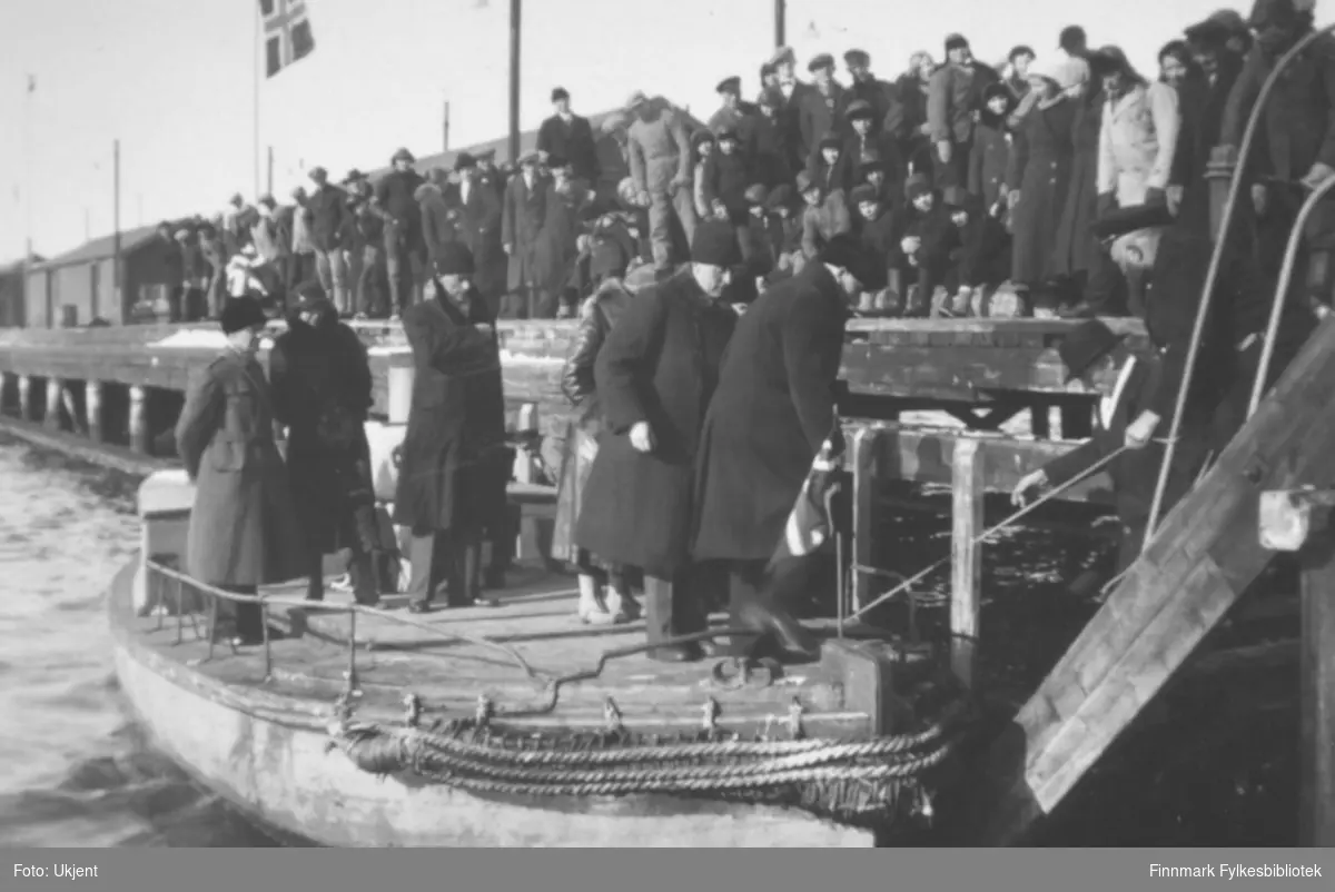 "Fra Dampskipskaia i Vadsø. Bildet viser et følge med bl.a kronprins Olav ombord i en liten båt i ferd med å gå i land. Kronprinsparet besøkte Finnmarksvidda påsken 1934. På baksiden av fotografiet står det skrevet: 'De kommer tilbake efter turen på havnen'.