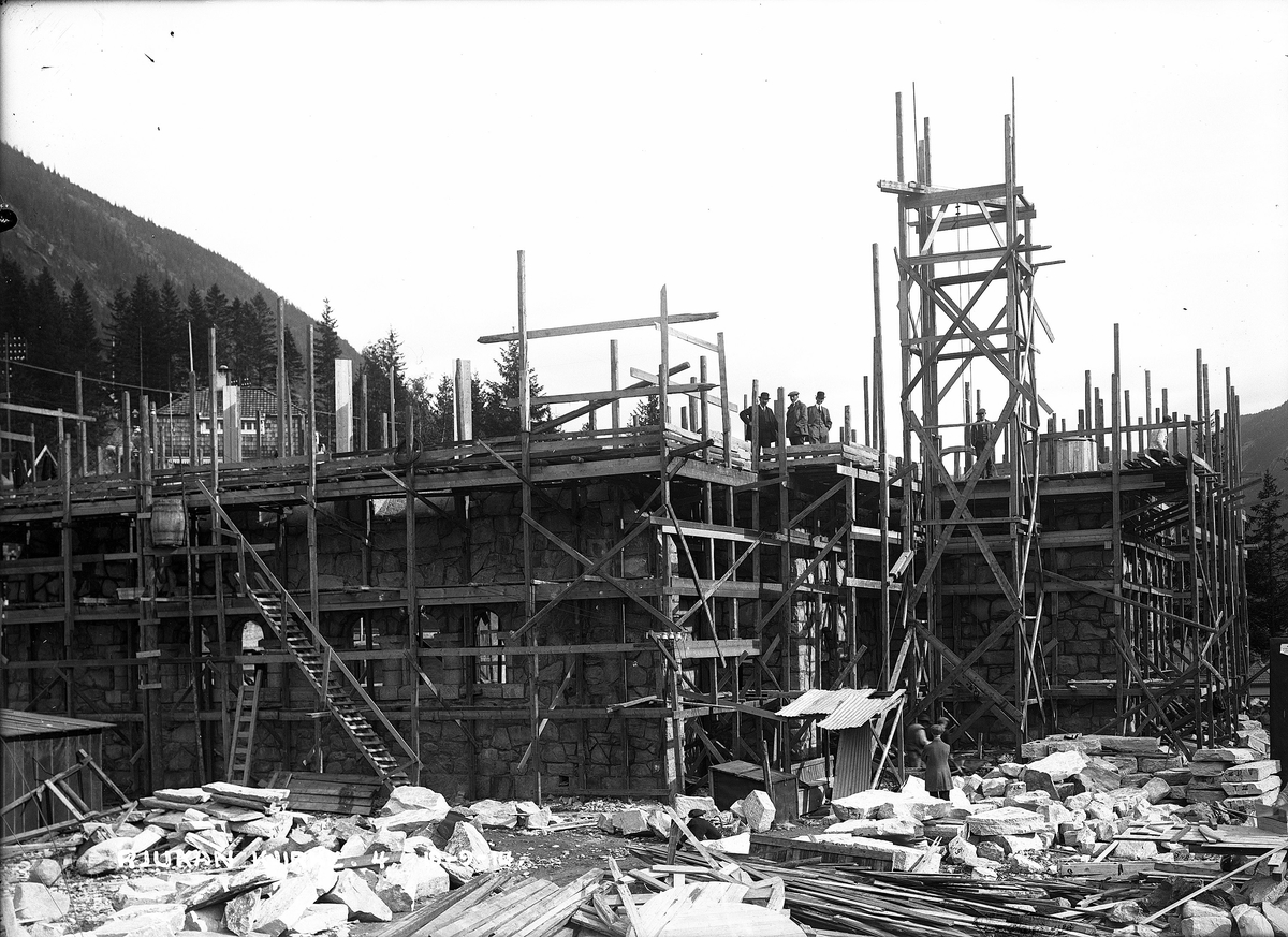 Rjukan kirke under bygging. Menn, antagelig ingeniører, stående på byggverkets andre etasje og en nede på bakkenivå. Naturstein til bruk på kirken liggende rundt på byggeplassen. Trestillas bygget rundt kirken.