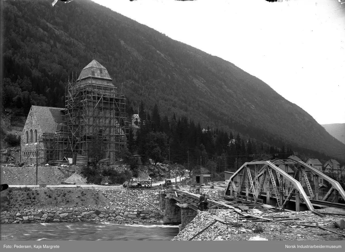 Byggearbeider på tårnet til Rjukan kirke. Trestillas rundt tårnet, bygningsmatrialer og skur stående på byggeplassen. Bro bygges over til Mæland fra Sam Eydes gate ved siden av jernbanebroen. Arbeidere stående ved broen.