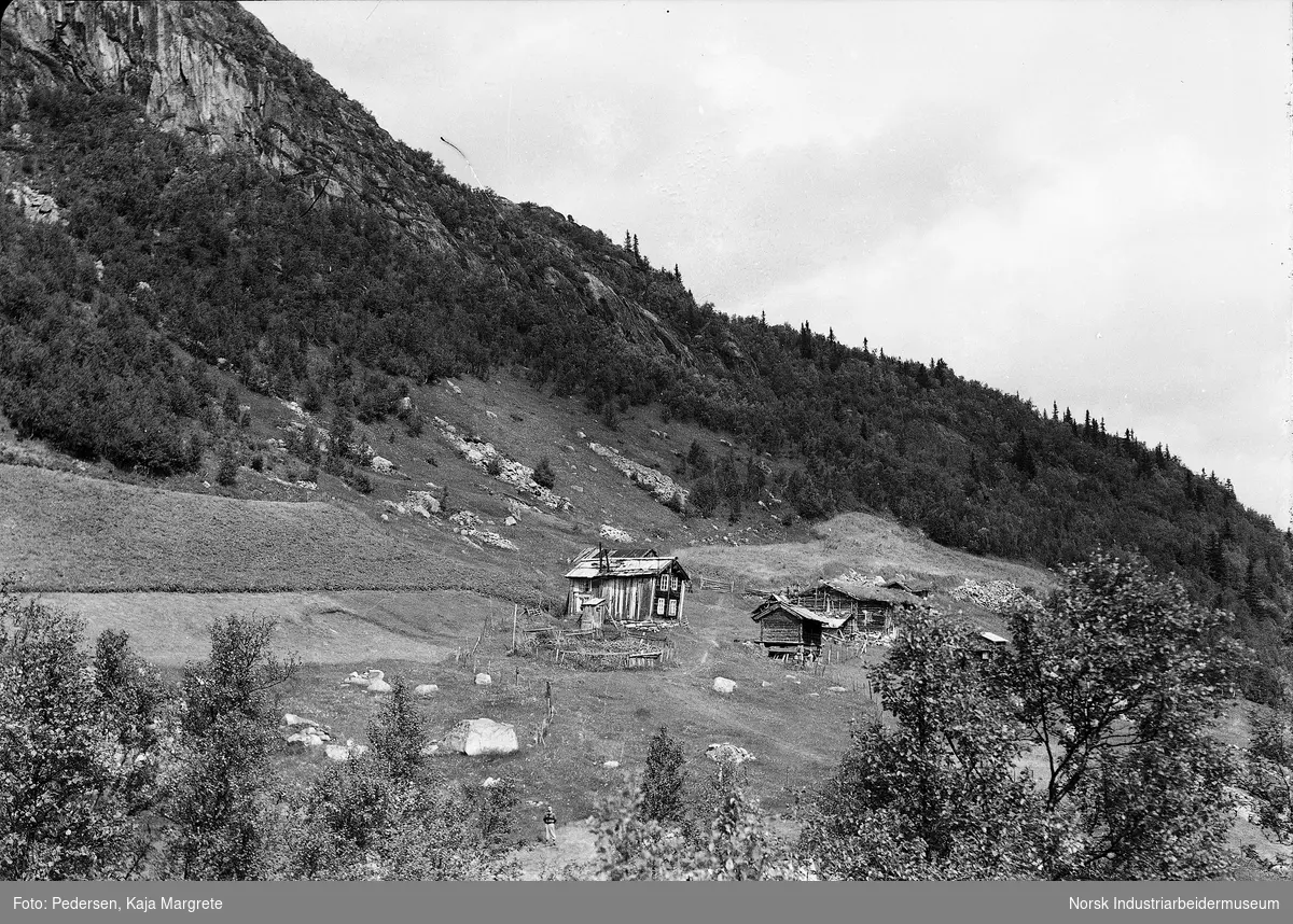 Berge fjellgård med seks bygninger. Menneske stående i forgrunnen.