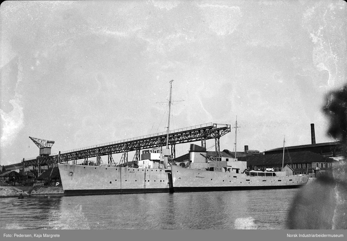 Båter, jageren Æger, sjøsatt 25. august 1936 og oppsynsskipet Nordkapp, sjøsatt 18. august 1937 liggende ved Marinens Hovedverft, Horten. Bildet er tatt i tiden etter sjøsettingen av oppsynsskipet Nordkapp og fartøyene ligger fortøyd ved verftet for utrustning, like ved beddingen. Bak fartøyene sees den såkalte "høybanen" med kran som løper langsmed beddingen.