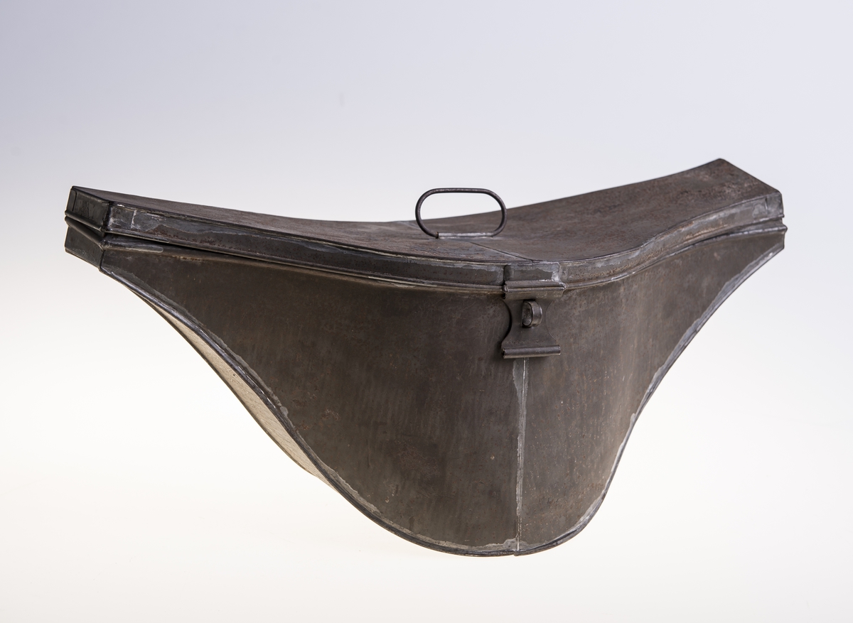 Metall hatteeske med hengslet lokk. Håndtak på lokket og en hempe på siden for hengelås. Esken har ikke fôr.
Hatten i silkefloss er tosnutet med gullbånddekor på hver snute. På den ene siden er det en kokarde med norske farger. Over denne ligger det gullmetallsnorer festet til en messingfarget knapp med motiv, den norske løve. Den ene side på hatten er høyere enn den andre. Hatten har fôr og svettebånd. Det er stempel inne i pullen.