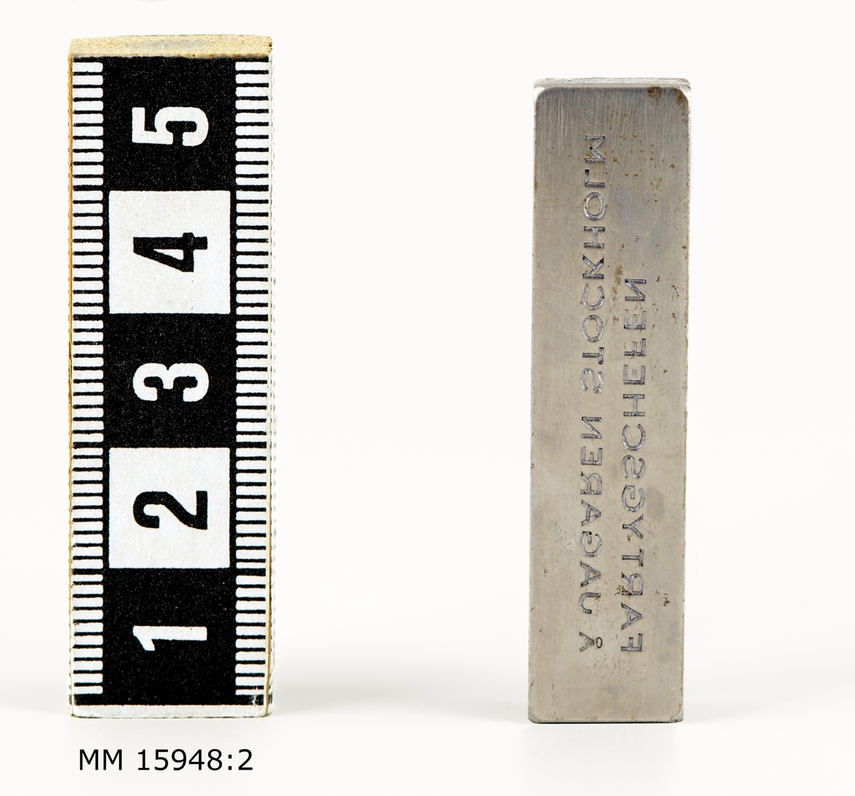 Rektangulärt metallstycke vars bredsida är inristad med texten: "FARTYGSCHEFEN Å JAGAREN STOCKHOLM".
Texten ses spegelvänt för betraktaren. Ovansidans kanter fasade.