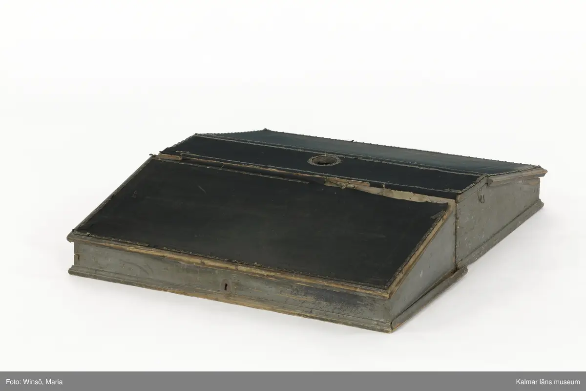 KLM 14272. Pulpet, kontorspulpet. Av trä, är på utsidan målad i grå färg. Baksidan omålad. Snedställt, uppfällbart lock klätt med fastnitat mörkt ylletyg. Nitar av mässing, i nitraderna sitter ett skinnliknande material. På framsidan ett nyckelhål med ett brett beslag av järn. På insidan ett inbyggt lås. Längst upp på pulpeten ett plant utrymme, i mitten ett hål, troligen för bläckhorn. Inuti, längst in, en hylla med fyra fack i två rader. På vänstra utsidan en krok, på den högra en ögla av metall. Har använts tillsammans med pulpet KLM 14273.