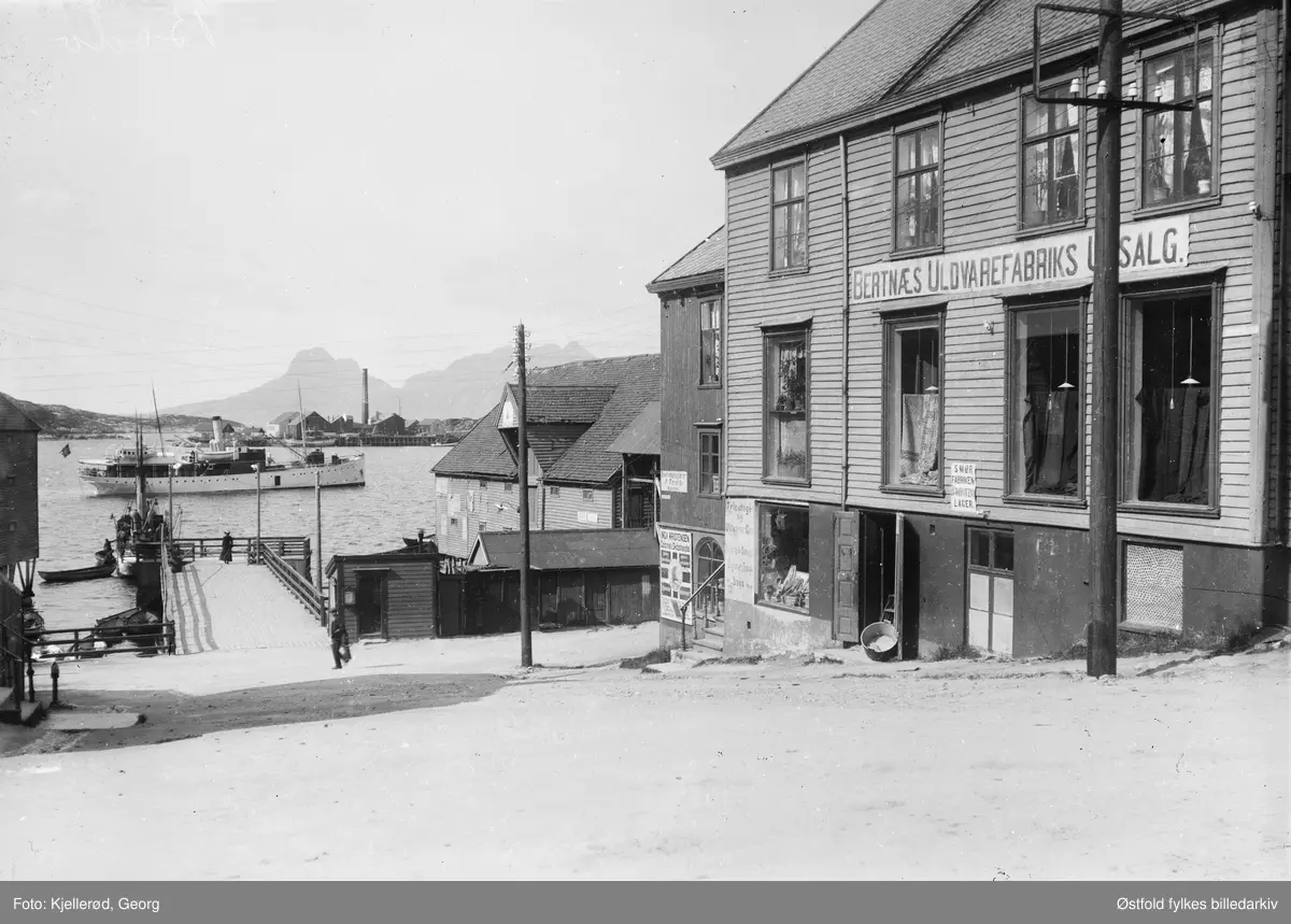 Gateparti fra Bodø med Bertnæs Uldvarefabrik udsalg til høyre.
Brygge med dampskip  midt i bildet.
Smørfabriken BJørgevins lager. 
Ingv. Kristensen Colonial og  Skipshandel.