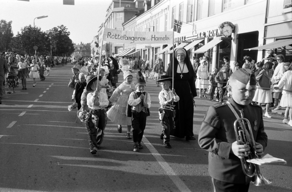 Diverse foto fra Barnas dag. Opptog i byen og aktiviteter på Hamar stadion. Barn i kostymer går i tog i Strandgata, Hamar. Barna bærer et banner med teksten "Rottefangeren fra Hammeln". En nonne følger barna, St. Torfinn barnehage. Mye folk i gata.