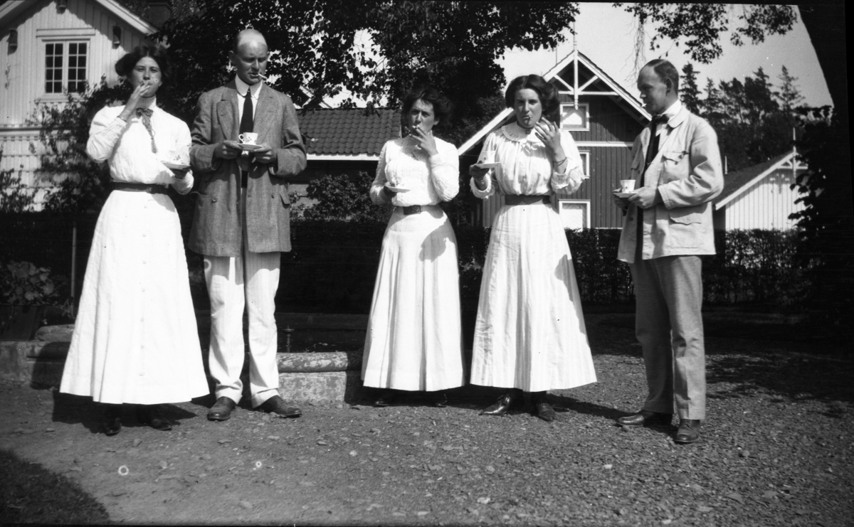 Fotoarkivet etter Gunnar Knudsen. Tre kvinner og to menn, som drikker kaffe og røyker. Bildet er tatt i haven på Borgestad gård. Til venstre ses Gunnar Knudsens eldste sønn, Erik Cappelen Knudsen. Mannen til høyre er antakelig Eriks bror Christen ("Kit") K. (1884-1986)