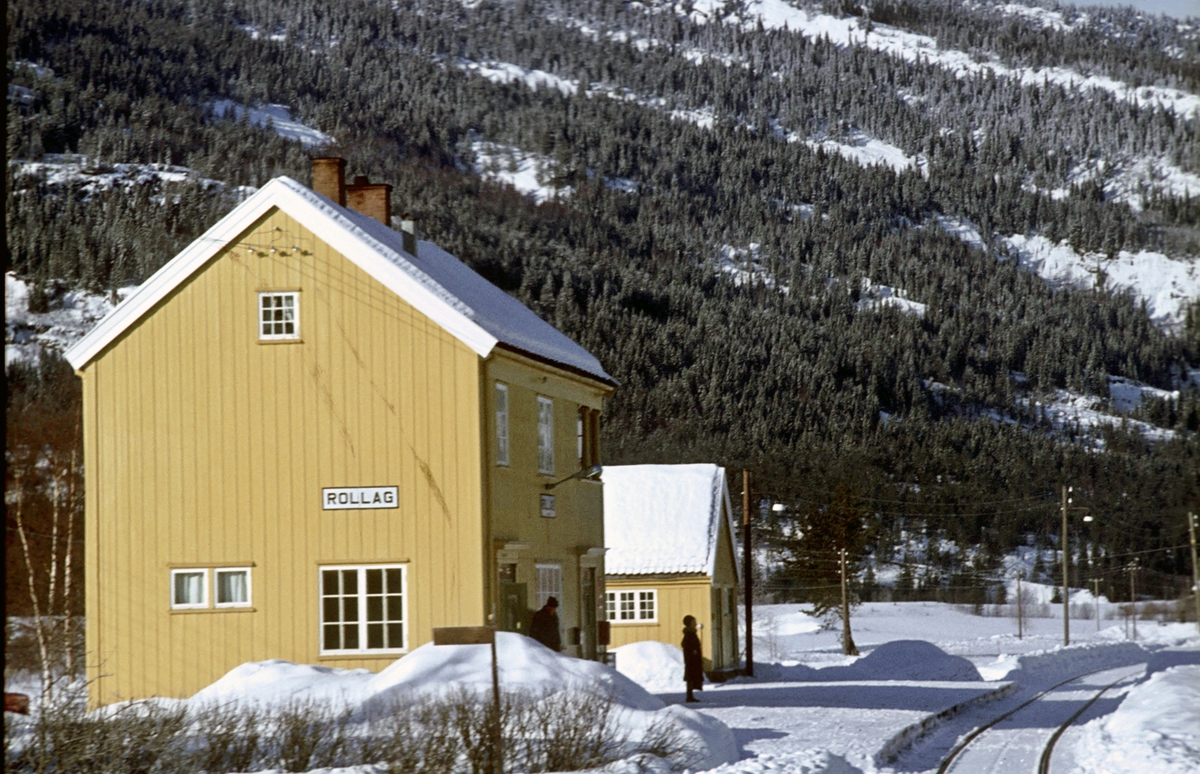 Rollag stasjon, Numedalsbanen. Numedal. Stasjonsbygning og uthus sett fra tog retning Rødberg. En reisende venter på toget.