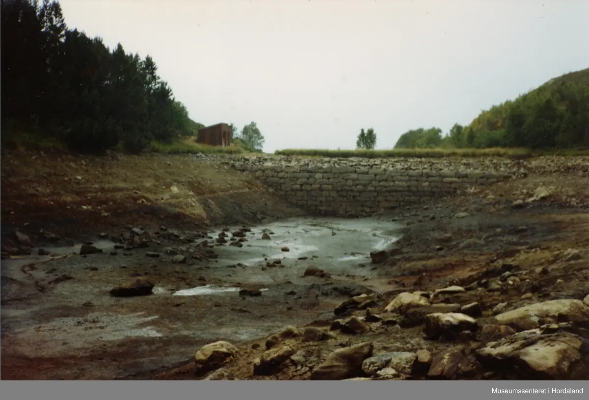Milastemma, demning i Salhus, Bergen kommune. Demning bygd til Salhus Tricotagefabrik (1859-1989), oppkalla etter disponent Emil Clausen, og bygd for å sikre vassforsyning til trikotasjefabrikken. Fotografiet viser stemma i ein tørkeperiode i 1982.