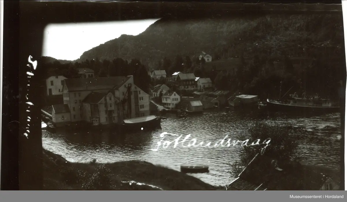 Bilde av Fotlandsvåg. Stort bedriftsbygg med små bygg rundt. Stor båt til høyre i bildet, og en robåt fremst i bildet.