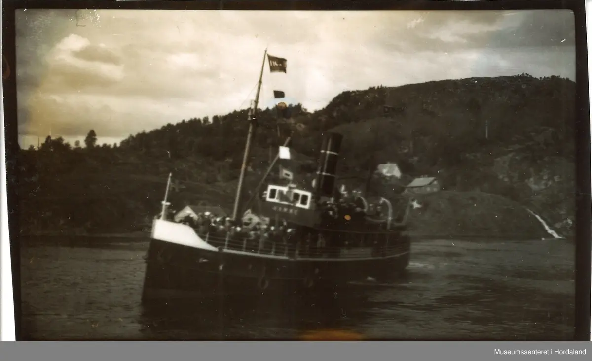 Dampskipet "Hamre" med masse folk. Norskflagg på båten, fjellandskap med små hus i bakgrunnen. Bildet er tatt fra kommunekaien i Fotlandsvåg.