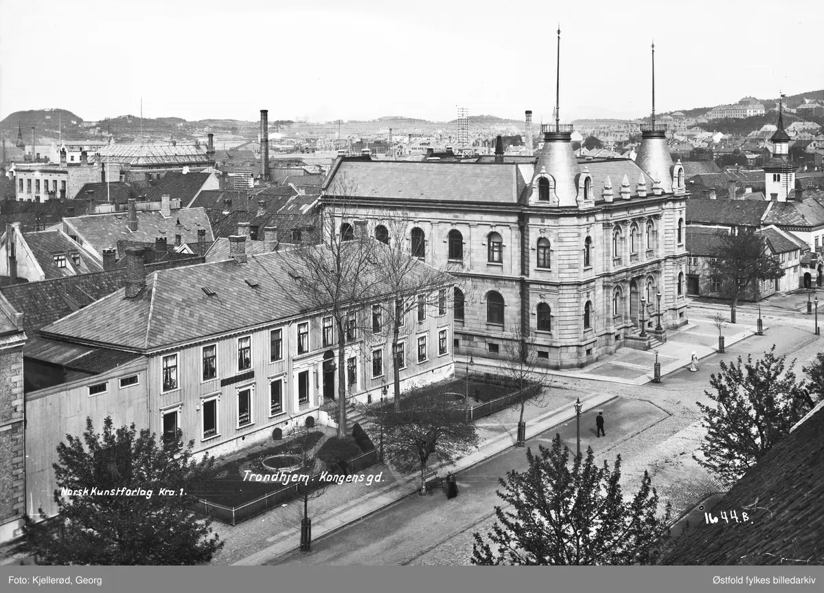 Gateparti fra Kongens gate i Trondheim. 

Fra høyre mot venstre: Gamle rådhus (med tårn), nå Folkebiblioteket. 2 små trehus, revet, nå brannstasjon. Søndre gate. Sparebanken. Apotekerveita. Løveapoteket, revet, nå Mercursenteret. Liten snipp av børs/ telegraf/ post.

"Bildet må være tatt fra nedre del av tårnet på Vår Frue kirke, noe av kirketaket i høyre billedkant.
I  venstre bildekant litt av den gamle Børs- og telegrafbygingen, deretter gammel apokergård (revet ca. slutten av 30-tallet for oppføring av ny telegrafbygning). Begge telegrafbygningene inngår i dag i kjøpesenteret Mercur). 
Apotekerveita går ned til venstre før Trondhjems Sparebank - byens  første bygning som ble oppført i stein etter middelalderen. Etter den går Søndre gate ned t.v. etterfulgt av den lave gamle brannstasjonen (erstattet 1942-1950 av ny, som i 2015 er under utflytting). 

Endelig en litt høyere bygning med tårn: Byens rådhus frem til ca. 1930, siden da Trondheim Folkebibliotek. 

Lenger bak, helt til venstre, høy bygning med flatt tak: Nordenfjeldske Kunstindustrimuseum (som senere fikk høyt saltak). Den høyeste fabrikkpipen mellom museet og banken tilhører Trondhjems Bryggeri, Dronningens gate 2. Høydedragene i bakgrunnen venstre side ligger på Ladehalvøya, ytterst t.v. Ladehammeren  Skogvokste høydedrag høyre side er Kuhaugen og (nærmest) Småbergan."
(Ivar Stav, 2015).