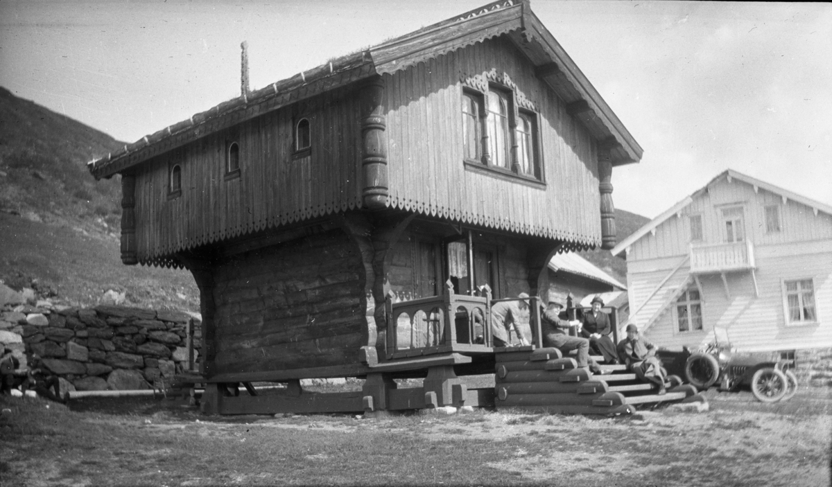 Fotoarkivet etter Gunnar Knudsen. Mennesker som sitter på trappen til et stabbur (bur) på Haukeliseter. Ved siden av stabburet står en automobil som ble brukt som rutebil. Bilen tilhører automobillaget Haukeli. Bildet er tatt i ca. 1910