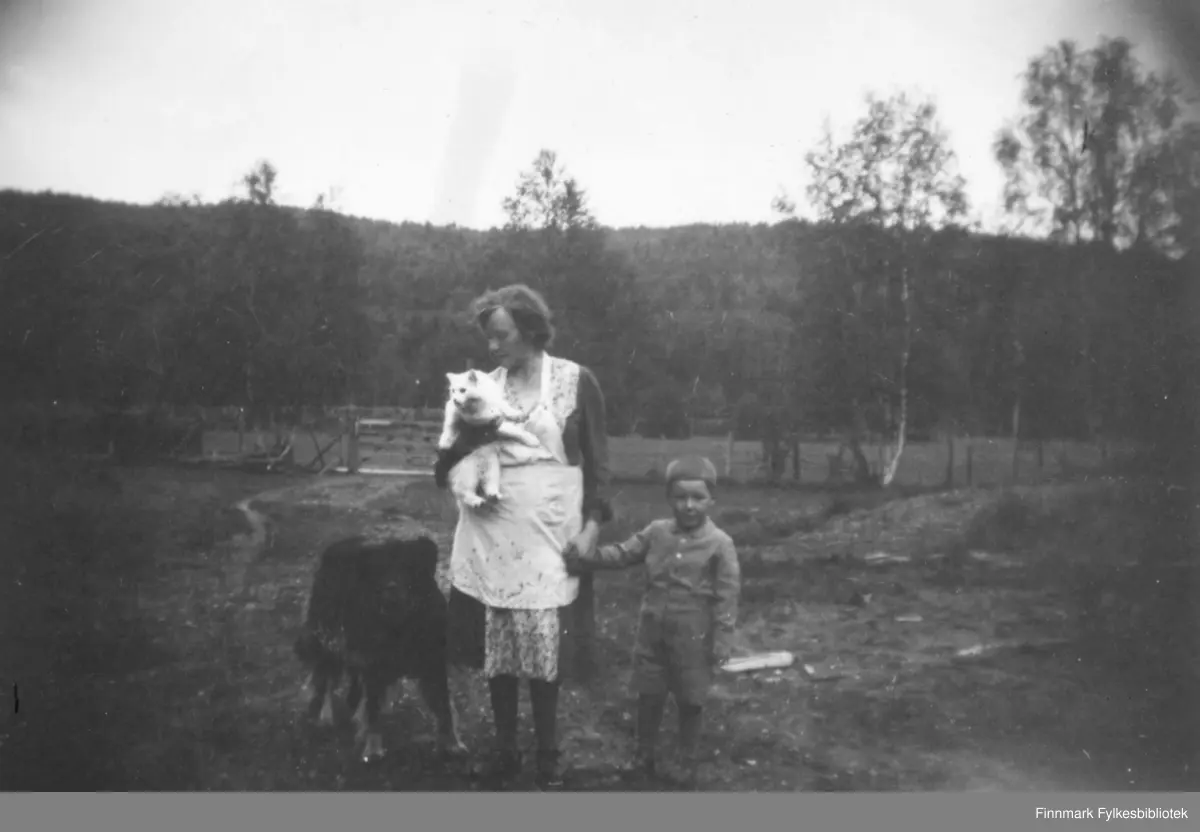 Magna Wisløff leier sin sønn Ole Arve Wisløff i hånden. Hun holder familiens katt og til venstre for henne står Kristian Wisløffs hund. Bak dem ser vi et gjerde og et jorde. I bakgrunnen tett skog. Ca.1945-50.