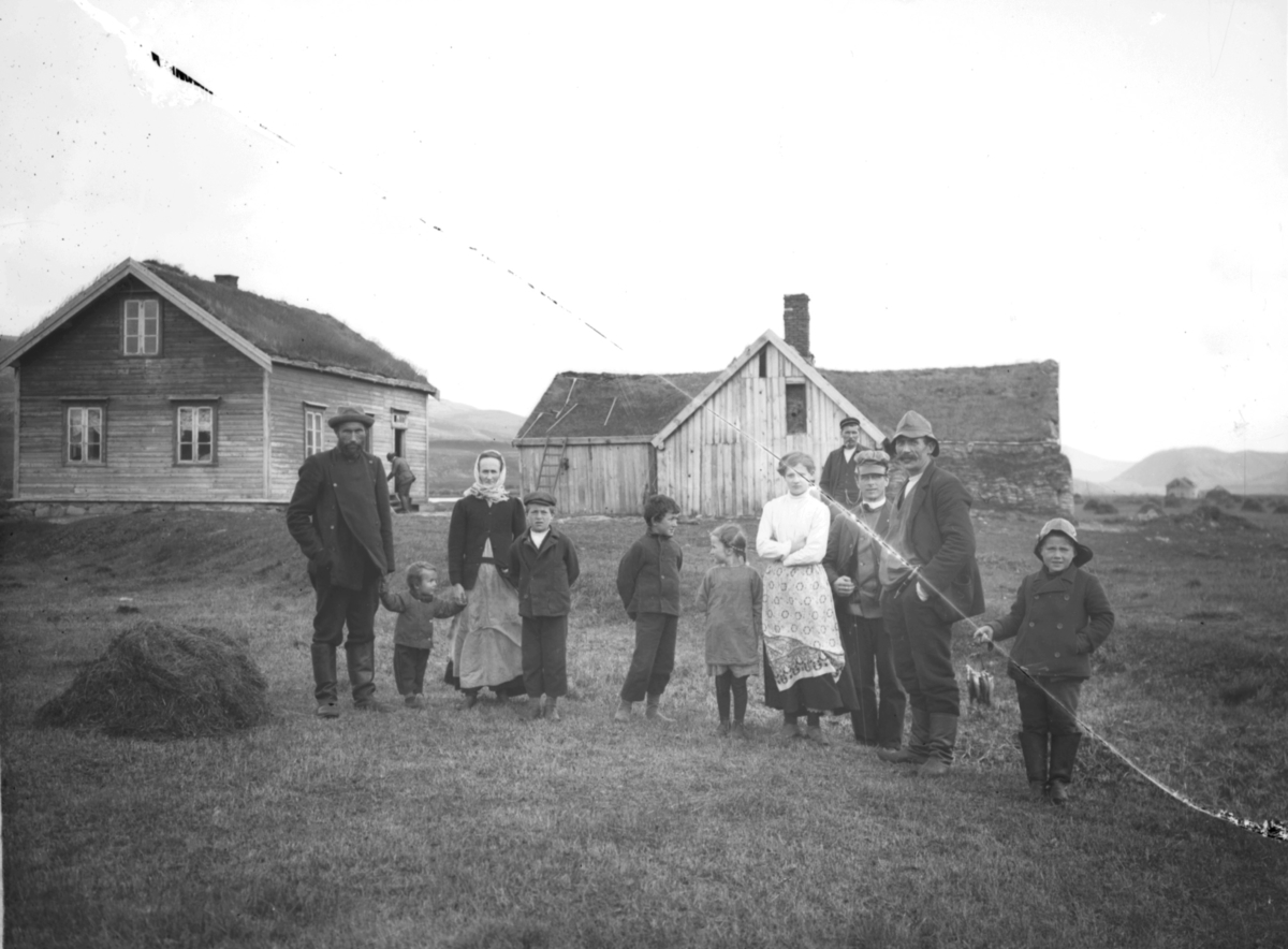 Gruppebilde på slåttemark i onnetida. Bolighus med uthus i bakgrunnen. Barn og voksne samlet. Grasmo er nr. 3 fra høyre. En gutt helt til høyre har på seg sydvest og holder en hanke småfisk i hånda. Ved våningshuset står en mann krokbøyd ved inngangsdøra. Høy rakt sammen i en haug til venstre i bildet