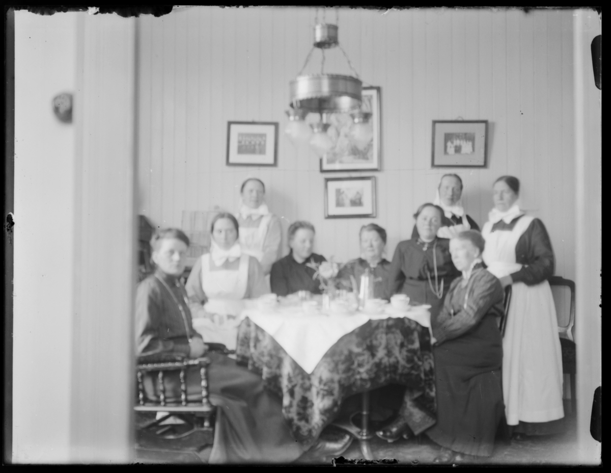 Kvinneforening holder møte i prestegården. Ni kvinner sitter og står rundt bordet. Noen er diakonisser i uniform. Nr. 4 fra venstre er fru Grasmo, Anne Marie Grasmo. Foran til høyre er Anne Gravem. (uskarp bilde, se bedre versjon FBib.82011-145)