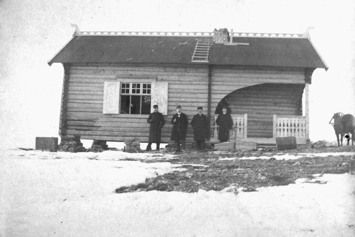 Bildet er trolig tatt på begynnelsen av 1900-tallet. På bildet står fire menn foran en hytte som antas å være skihytten ved basis A i Vadsø. Mennene er kledd i frakker, bukser og hatter. Enkelte av mennene har bart og holder muligens en pipe eller tobakk. På bygget kan man se et stort vindu og en inngang med gelender. På taket er det blitt laget dekorativ pynt, skorstein og en stige. Ved siden av bygget kan man se en hest med utstyr. Under bygget er det blitt stablet steiner. Foran gelenderet ligger det en tønne. Bakken er dekket til med snø.