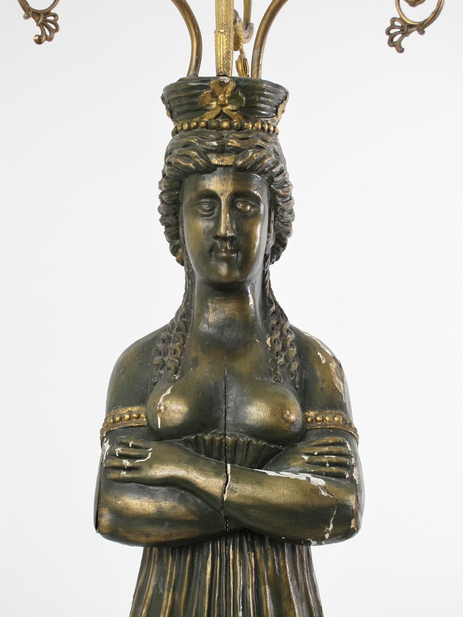 Kandelaber med karyatide. Mahogny, bronsert tre, messingblikk messing. Søylefot med mahogny sokkel i to trinn, det nedre med en  list rundt  i lysere tre. Dorisk søyle, bronsert med  forgylt  kapitel. Over dette en mahognysokkel i to trinn som base  for karyatide i bronsert tre på firkantet liten fotstykke. Karyatiden er helt frontal og symmetrisk, med korslagte armer.  Fotsidt draperi med vertikale folder, som lar bryst og skuldre være  bare.  I ryggen har draperiet folder som faller i bølger tvers over  kroppen, og det holdes oppe av et belte midt over ryggen. Om bakhodet et bånd og  rutenett.  på hodet en krone med utskrånende sider og rille  pyntet med bånd med trebladet blomst. Over kronen et beslag, støpt i messing, skrudd fast i figurens hode, med et spyd  med  ekenøtt midt i og  tre lysarmer.