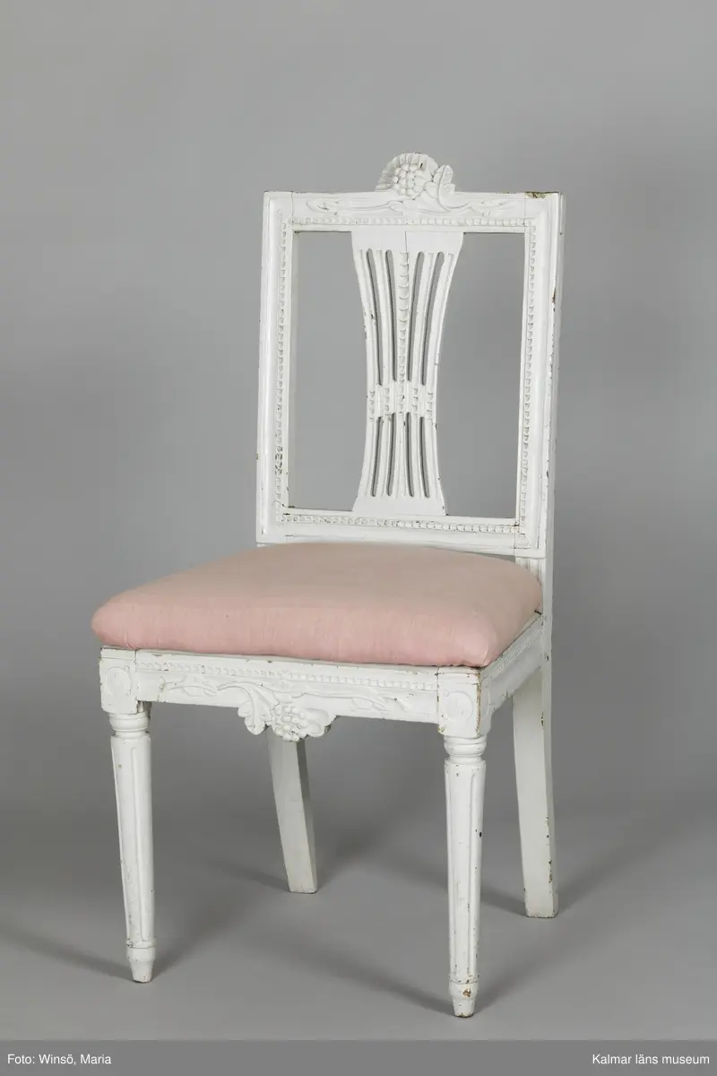 KLM 12727:2. Stol, ryggbrickstol av trä. 8 st. Två något olika grupper av stolar med fyra av varje, den ena gruppen med förgyllda detaljer och den andra helt vitmålad. I övrigt likadan dekor. Den ena gruppen stolar (stol 1-4) är vitmålade med guldfärgad äggstavsdekor på fram- och sidosargarna. Dekor på ryggöverstycke samt framsarg i form av humlekvistar. Benen är kannelerade, delvis förgyllda och nedåtavsmalnande. Ryggbrickan är genombruten och har en form av ett kopplat spjälknippe. Sitsen är stoppad och klädd med textil med ränder med blomsterslingor. Textilen är mycket blekt och originalfärgen syns på några ställen, då i grönt. Gruppen med stolar som är utan förgyllning (stol 5-8) har tidigare haft sitsarna klädda med rosa textil med mönster av lagerkransar med urna i mitten. Likadan textil kan ses på dyna i soffan KLM 27787. Gustaviansk.