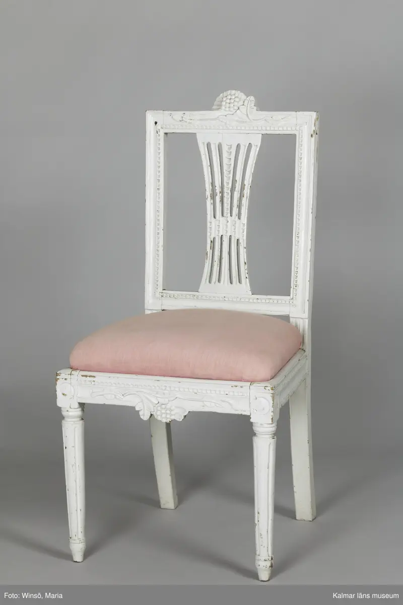 KLM 12727:2. Stol, ryggbrickstol av trä. 8 st. Två något olika grupper av stolar med fyra av varje, den ena gruppen med förgyllda detaljer och den andra helt vitmålad. I övrigt likadan dekor. Den ena gruppen stolar (stol 1-4) är vitmålade med guldfärgad äggstavsdekor på fram- och sidosargarna. Dekor på ryggöverstycke samt framsarg i form av humlekvistar. Benen är kannelerade, delvis förgyllda och nedåtavsmalnande. Ryggbrickan är genombruten och har en form av ett kopplat spjälknippe. Sitsen är stoppad och klädd med textil med ränder med blomsterslingor. Textilen är mycket blekt och originalfärgen syns på några ställen, då i grönt. Gruppen med stolar som är utan förgyllning (stol 5-8) har tidigare haft sitsarna klädda med rosa textil med mönster av lagerkransar med urna i mitten. Likadan textil kan ses på dyna i soffan KLM 27787. Gustaviansk.