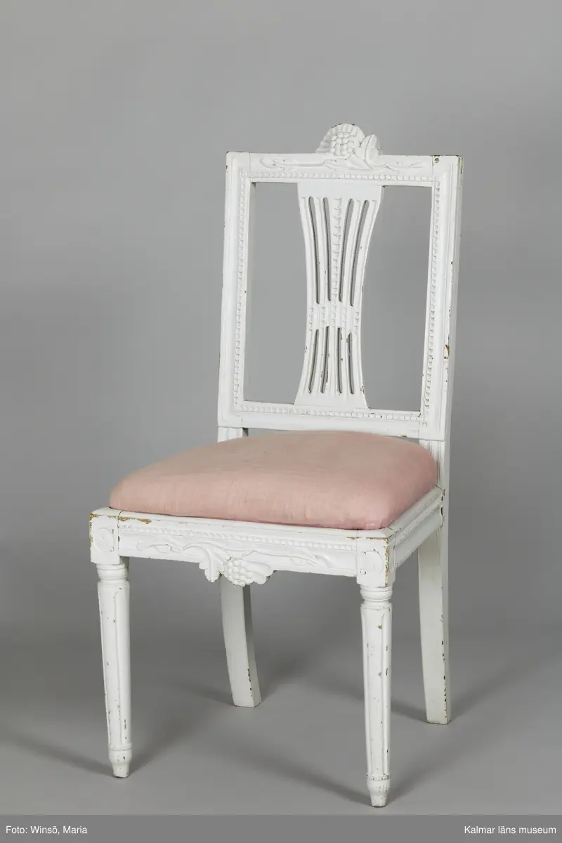 KLM 12727:2. Stol, ryggbrickstol av trä. 8 st. Två något olika grupper av stolar med fyra av varje, den ena gruppen med förgyllda detaljer och den andra helt vitmålad. I övrigt likadan dekor. Den ena gruppen stolar (stol 1-4) är vitmålade med guldfärgad äggstavsdekor på fram- och sidosargarna. Dekor på ryggöverstycke samt framsarg i form av humlekvistar. Benen är kannelerade, delvis förgyllda och nedåtavsmalnande. Ryggbrickan är genombruten och har en form av ett kopplat spjälknippe. Sitsen är stoppad och klädd med textil med ränder med blomsterslingor. Textilen är mycket blekt och originalfärgen syns på några ställen, då i grönt. Gruppen med stolar som är utan förgyllning (stol 5-8) har tidigare haft sitsarna klädda med rosa textil med mönster av lagerkransar med urna i mitten. Likadan textil kan ses på dyna i soffan KLM 27787. Gustaviansk.