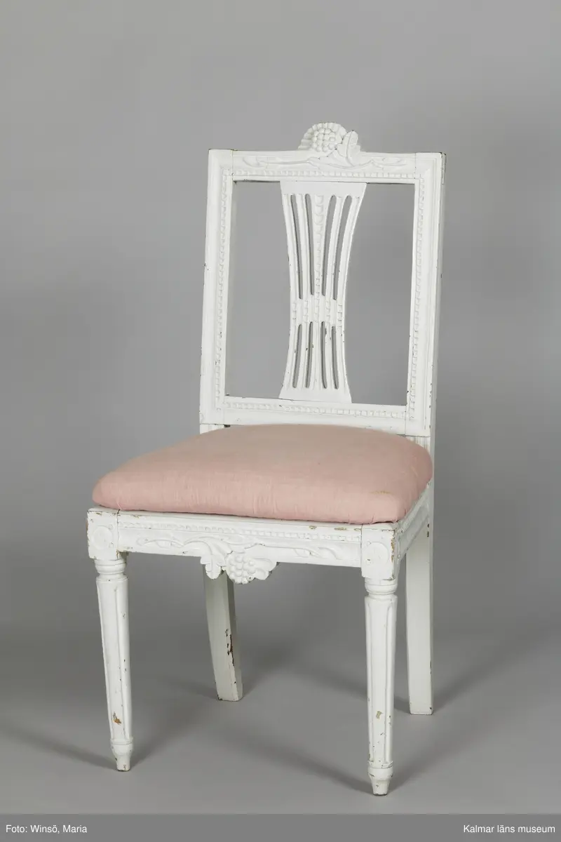 KLM 12727:2. Stol, ryggbrickstol av trä. 8 st. Två något olika grupper av stolar med fyra av varje, den ena gruppen med förgyllda detaljer och den andra helt vitmålad. I övrigt likadan dekor. Den ena gruppen stolar (stol 1-4) är vitmålade med guldfärgad äggstavsdekor på fram- och sidosargarna. Dekor på ryggöverstycke samt framsarg i form av humlekvistar. Benen är kannelerade, delvis förgyllda och nedåtavsmalnande. Ryggbrickan är genombruten och har en form av ett kopplat spjälknippe. Sitsen är stoppad och klädd med textil med ränder med blomsterslingor. Textilen är mycket blekt och originalfärgen syns på några ställen, då i grönt. Gruppen med stolar som är utan förgyllning (stol 5-8) har tidigare haft sitsarna klädda med rosa textil med mönster av lagerkransar med urna i mitten. Likadan textil kan ses på dyna i soffan KLM 27787. Gustaviansk.