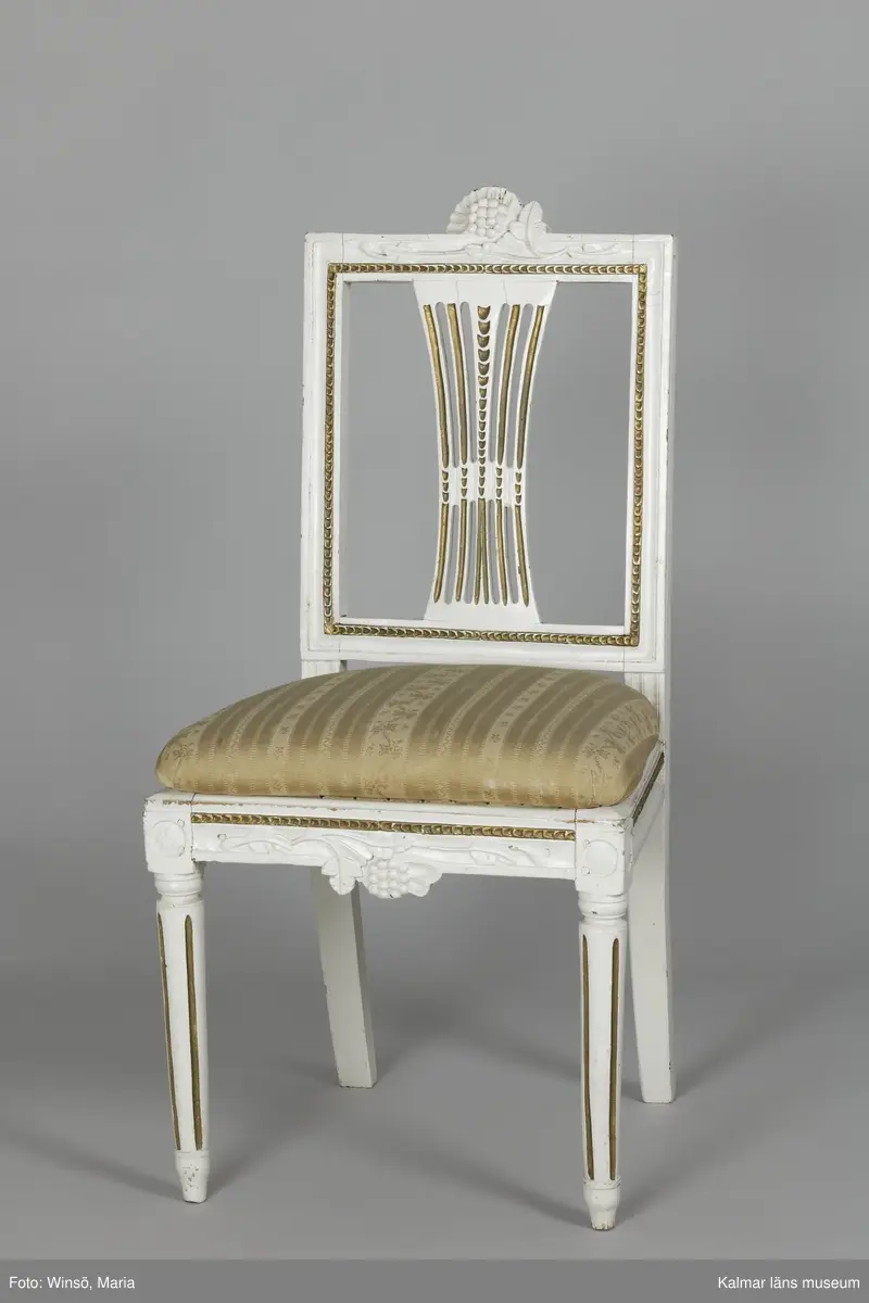 KLM 12727:2. Stol, ryggbrickstol av trä. 8 st. Två något olika grupper av stolar med fyra av varje, den ena gruppen med förgyllda detaljer och den andra helt vitmålad. I övrigt likadan dekor. Den ena gruppen stolar (stol 1-4) är vitmålade med guldfärgad äggstavsdekor på fram- och sidosargarna. Dekor på ryggöverstycke samt framsarg i form av humlekvistar. Benen är kannelerade, delvis förgyllda och nedåtavsmalnande. Ryggbrickan är genombruten och har en form av ett kopplat spjälknippe. Sitsen är stoppad och klädd med textil med ränder med blomsterslingor. Textilen är mycket blekt och originalfärgen syns på några ställen, då i grönt. Gruppen med stolar som är utan förgyllning (stol 5-8) har tidigare haft sitsarna klädda med rosa textil med mönster av lagerkransar med urna i mitten. Likadan textil kan ses på dyna i soffan KLM 27787. Gustaviansk.