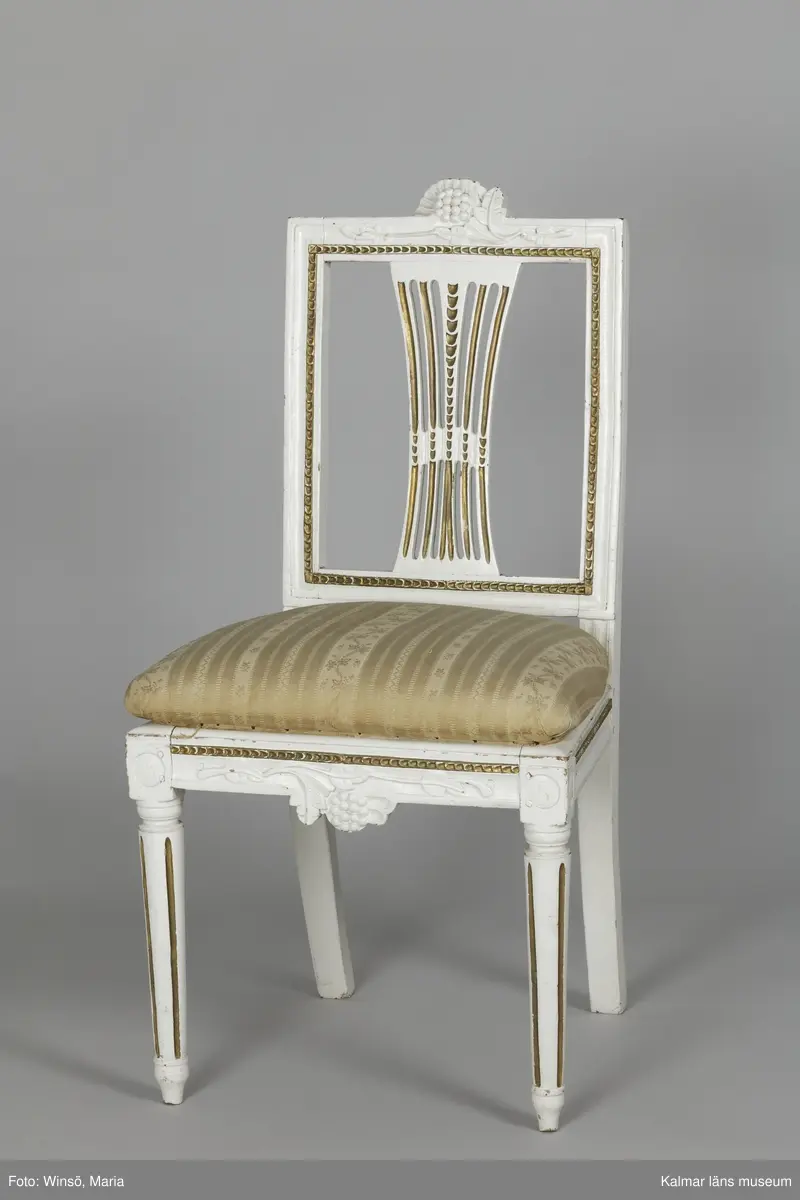 KLM 12727:2. Stol, ryggbrickstol av trä. 8 st. Två något olika grupper av stolar med fyra av varje, den ena gruppen med förgyllda detaljer och den andra helt vitmålad. I övrigt likadan dekor. Den ena gruppen stolar (stol 1-4) är vitmålade med guldfärgad äggstavsdekor på fram- och sidosargarna. Dekor på ryggöverstycke samt framsarg i form av humlekvistar. Benen är kannelerade, delvis förgyllda och nedåtavsmalnande. Ryggbrickan är genombruten och har en form av ett kopplat spjälknippe. Sitsen är stoppad och klädd med textil med ränder med blomsterslingor. Textilen är mycket blekt och originalfärgen syns på några ställen, då i grönt. Gruppen med stolar som är utan förgyllning (stol 5-8) har tidigare haft sitsarna klädda med rosa textil med mönster av lagerkransar med urna i mitten. Likadan textil kan ses på dyna i soffan KLM 27787. Gustaviansk.