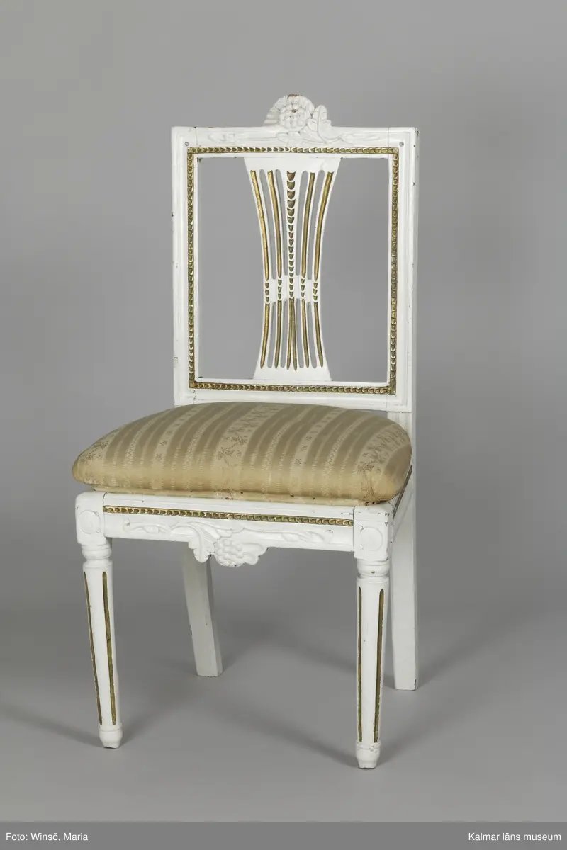 KLM 12727:2. Stol, ryggbrickstol av trä. 8 st. Två något olika grupper av stolar med fyra av varje, den ena gruppen med förgyllda detaljer och den andra helt vitmålad. I övrigt likadan dekor. Den ena gruppen stolar (stol 1-4) är vitmålade med guldfärgad äggstavsdekor på fram- och sidosargarna. Dekor på ryggöverstycke samt framsarg i form av humlekvistar. Benen är kannelerade, delvis förgyllda och nedåtavsmalnande. Ryggbrickan är genombruten och har en form av ett kopplat spjälknippe. Sitsen är stoppad och klädd med textil med ränder med blomsterslingor. Textilen är mycket blekt och originalfärgen syns på några ställen, då i grönt. Gruppen med stolar som är utan förgyllning (stol 5-8) har tidigare haft sitsarna klädda med rosa textil med mönster av lagerkransar med urna i mitten. Likadan textil kan ses på dyna i soffan KLM 27787. Gustaviansk.