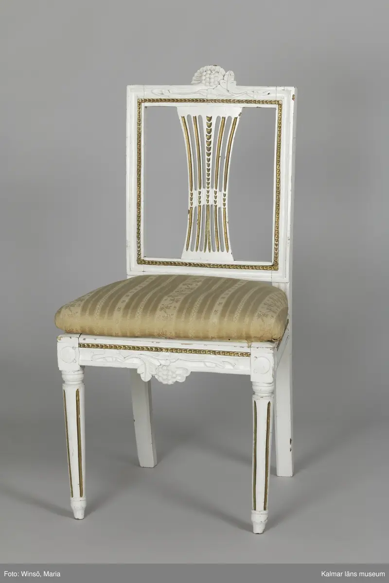 KLM 12727:2. Stol, ryggbrickstol av trä. 8 st. Två något olika grupper av stolar med fyra av varje, den ena gruppen med förgyllda detaljer och den andra helt vitmålad. I övrigt likadan dekor. Den ena gruppen stolar (stol 1-4) är vitmålade med guldfärgad äggstavsdekor på fram- och sidosargarna. Dekor på ryggöverstycke samt framsarg i form av humlekvistar. Benen är kannelerade, delvis förgyllda och nedåtavsmalnande. Ryggbrickan är genombruten och har en form av ett kopplat spjälknippe. Sitsen är stoppad och klädd med textil med ränder med blomsterslingor. Textilen är mycket blekt och originalfärgen syns på några ställen, då i grönt. Gruppen med stolar som är utan förgyllning (stol 5-8) har tidigare haft sitsarna klädda med rosa textil med mönster av lagerkransar med urna i mitten. Likadan textil kan ses på dyna i soffan KLM 27787. Gustaviansk.