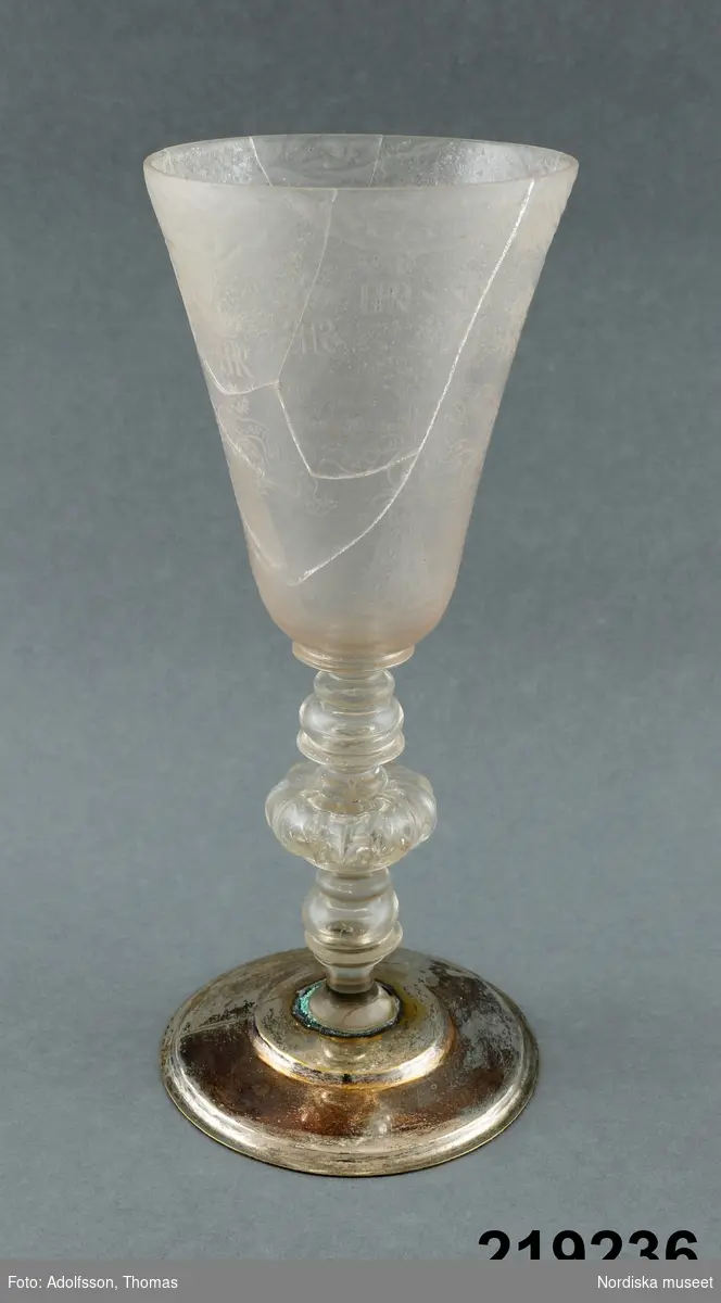 Huvudliggaren:
"Pokal, glas, graverat; fot yngre, silver; franska och svenska inskrifter. Fredrik I:s namnchiffer o. andra konungars; Skattk. kat. 366; g. av okänd givare år 1877. Från Statens Hist. Museum, Stockholm 6102:b."

Bilaga:
Inget utöver huvudliggaren.

Katalogkort I:
"Pokal. Kuppa och ben av glas på silverfot. Graverad.
Inskription: Att Wänskap medh Frögd - kollas
Kring brädden draperidekor.
6 kungliga monogram: Fredrik I:s ( F och FRS), FRP= Fredericus Rex Russiae, GRB=Gregorius Rex Brittaniae, FRD= Fredericus Rex Daniae, PZM= Peter Tzar Museoviae.
Dekoren syftar på freden med Hannover och England i Sthlm 1719, med Preussen i Sthlm 1720, med Danmark på Fredriksborgs slott 1720, med Ryssland i Nystad 1721.
Dessutom fem lageromvirade pyramider med symboler och språkband.
Massa: mjölkaktigt opak. Korrosionprocess starkt framskriden.

katalogkort II:
"Graverad dekor. 

Se Skattkammarkatalogen 1930, nr 366
samt Seitz 1936, pl. 5