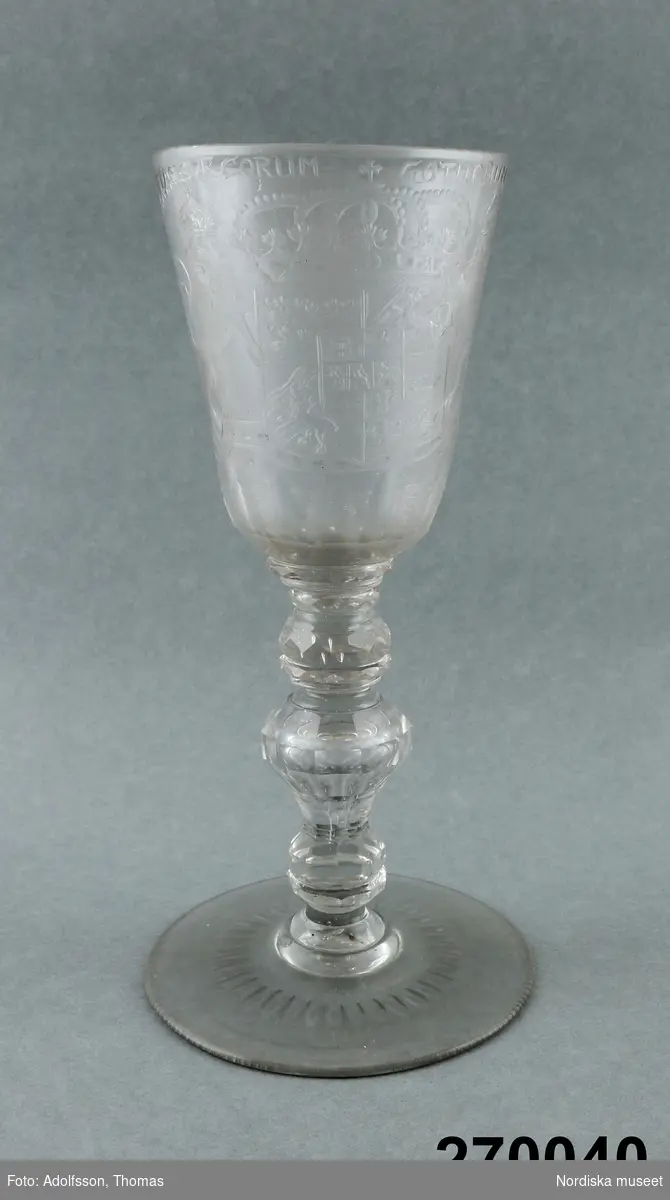 Huvudliggaren:
Pokal, glas; på rund fot, ben med tre vulster, slipad dekor, på kupan graverat svenska riksvapnet samt Hessen-Cassels vapen; inskr. 'FRIEDERICUS:DEI:GRATIAS:SVE:CORUM:GOTHORUM:VANDALORUM:QUE:REX'; på motsatta sidan sol; spår av glassjuka.  23,8 cm, Diam kupa 9,5 cm. Kungsholms glasbruk."

