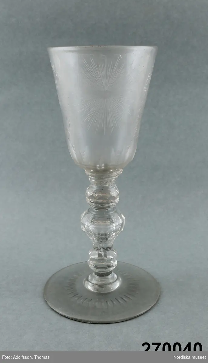 Huvudliggaren:
Pokal, glas; på rund fot, ben med tre vulster, slipad dekor, på kupan graverat svenska riksvapnet samt Hessen-Cassels vapen; inskr. 'FRIEDERICUS:DEI:GRATIAS:SVE:CORUM:GOTHORUM:VANDALORUM:QUE:REX'; på motsatta sidan sol; spår av glassjuka.  23,8 cm, Diam kupa 9,5 cm. Kungsholms glasbruk."
