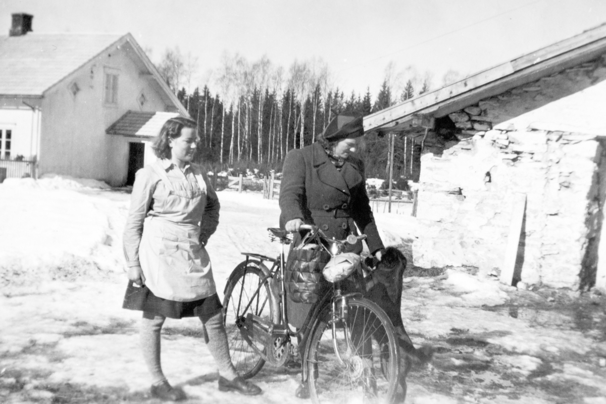 Marta H. Tomter og Karen E: Tomter (svigerinner) foran gammelfjøset på Søndre Tomter (Ner-Tomter) ca. 1950. Buhunden «Lita». Sykkel, veske og pakke på styret.