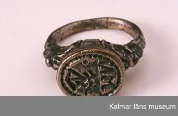 KLM 16729. Sigillring, av silver. Trind ring, oval tjock klack. Renässansornament i övergången mellan ring och klack. I klacken bokstäverna MN samt vapensköld med bomärke. Datering: 1500-tal eller tidigt 1600-tal.