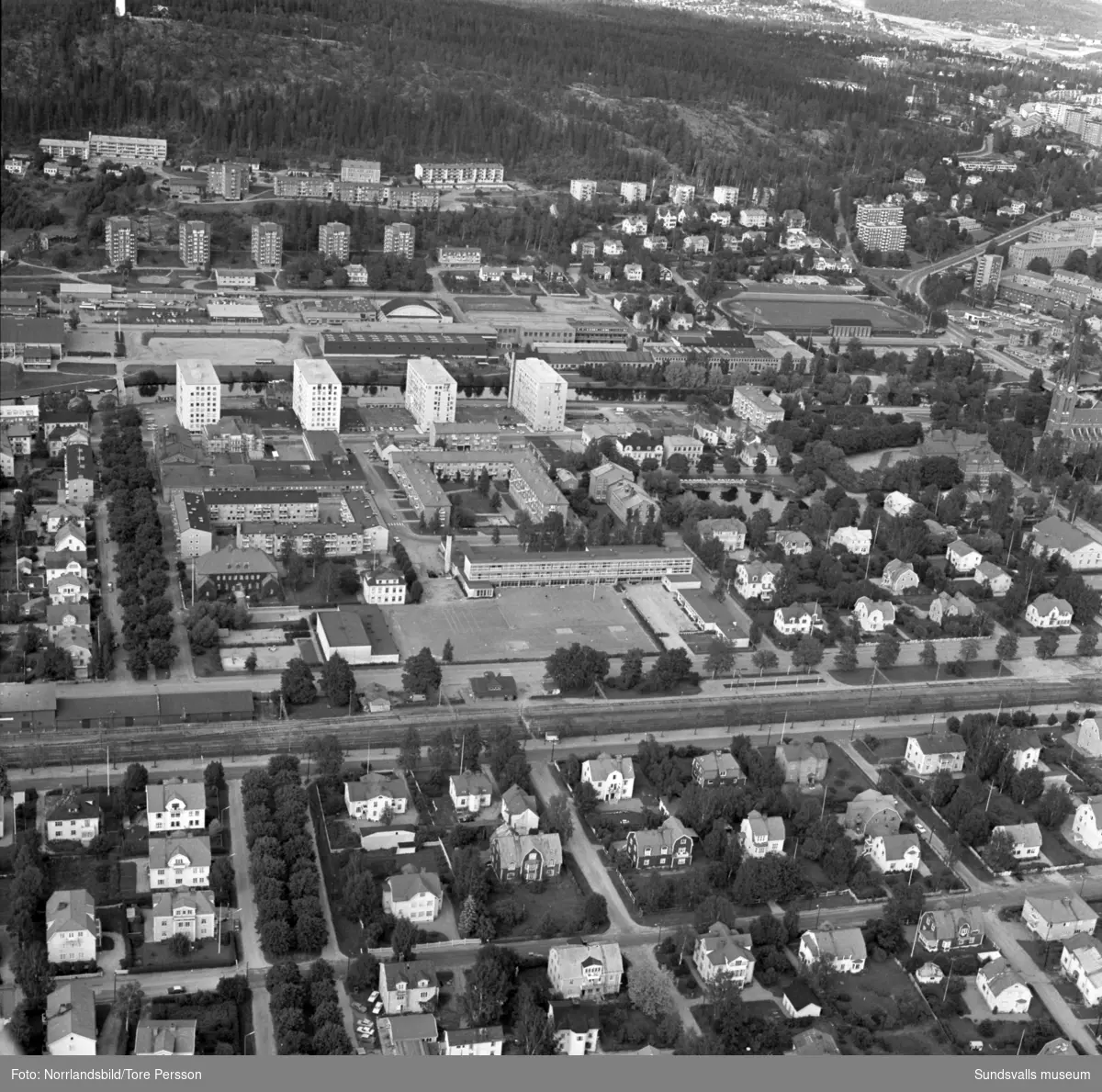 Flygfoto över västra delen av centrala Sundsvall. Från Västra Långgatan längst ner i bild via Åkersviks skola, Storgatan 52-58, Idrottsparken, Tennishallen, Sporthallen, Alliero och överst i bild syns utsiktstornet på Norra Berget.