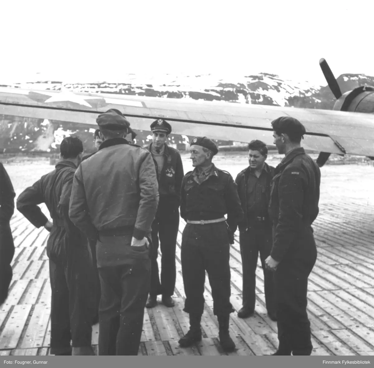 Lakselv. Frigjøringsvåren 1945. På det tyske plankedekket Banak flyplass står en gruppe menn foran et fly (Bernt Balchens Dakota DC-3), noen i militære uniformer. Kaptein Lunde i midten. Plankene fra flyplassen ble etter krigen gjenbrukt i mange bygninger i Lakselvdistriktet.
