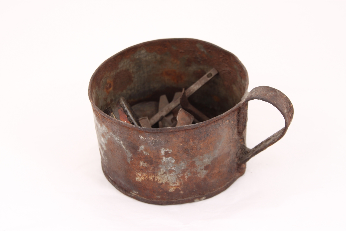 En metallkopp med ulike gjenstander. 
Koppen inneholder; en beltespenne, to runde metallbokser, tre pinsetter, diverse nøkler(?), tråd til beksøm(?) og skruer i ulik størrelse.