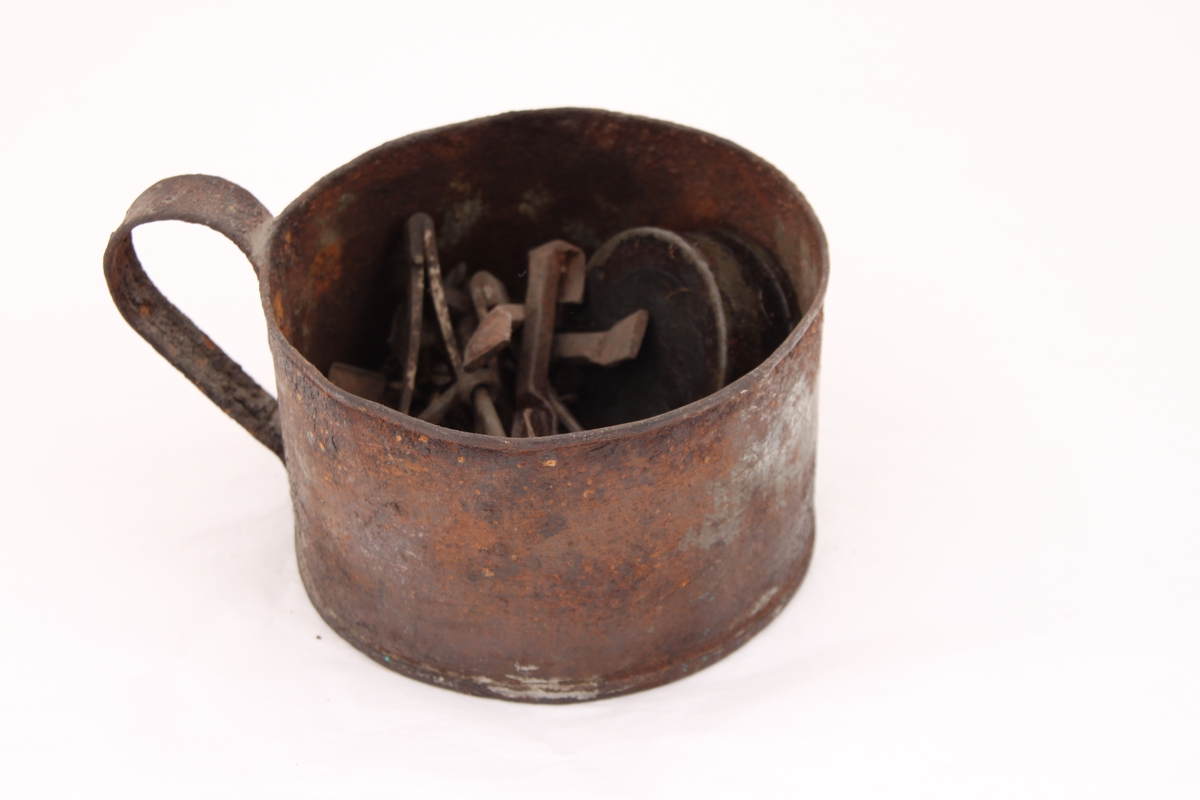 En metallkopp med ulike gjenstander. 
Koppen inneholder; en beltespenne, to runde metallbokser, tre pinsetter, diverse nøkler(?), tråd til beksøm(?) og skruer i ulik størrelse.