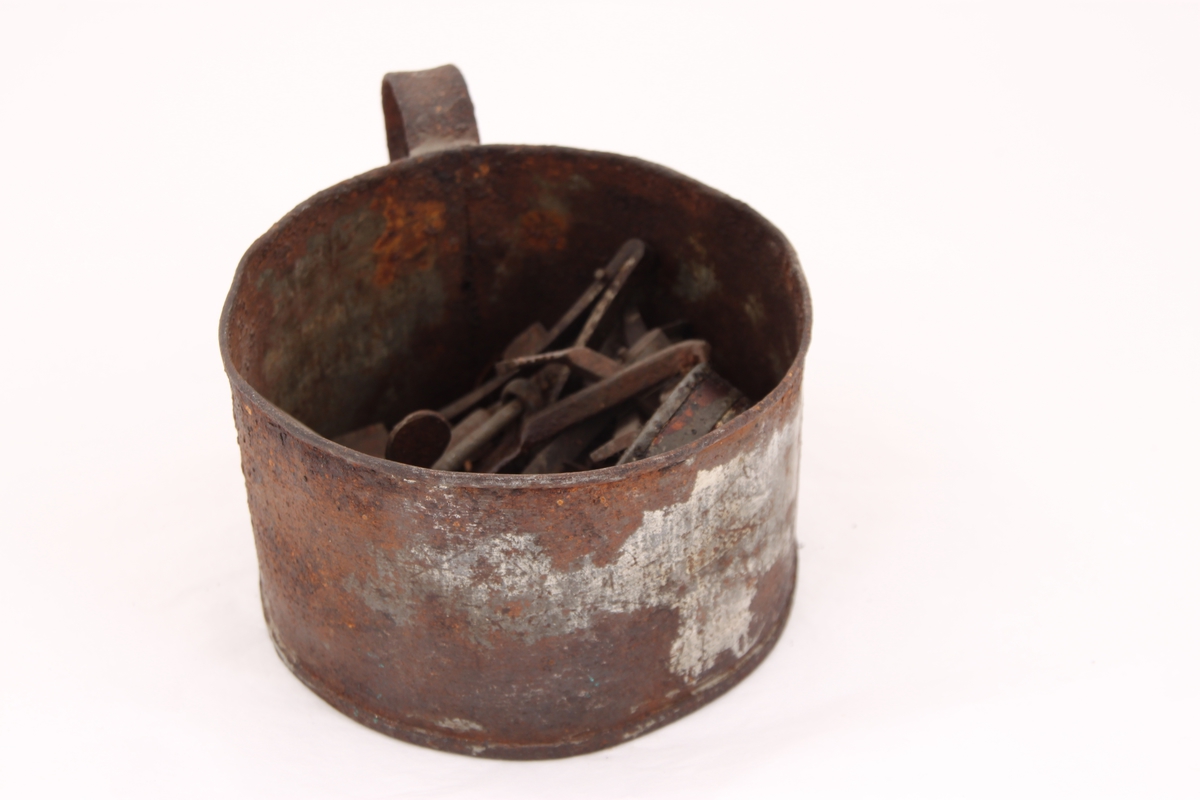 En metallkopp med ulike gjenstander. 
Koppen inneholder; en beltespenne, to runde metallbokser, tre pinsetter, diverse nøkler(?), tråd til beksøm(?) og skruer i ulik størrelse.