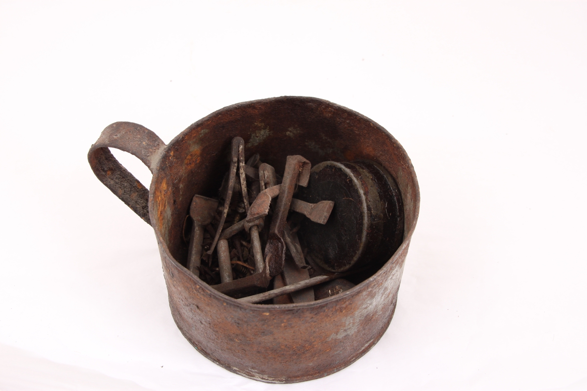 En metallkopp med ulike gjenstander. 
Koppen inneholder; en beltespenne, to runde metallbokser, tre pinsetter, diverse nøkler(?), tråd til beksøm(?) og skruer i ulik størrelse.