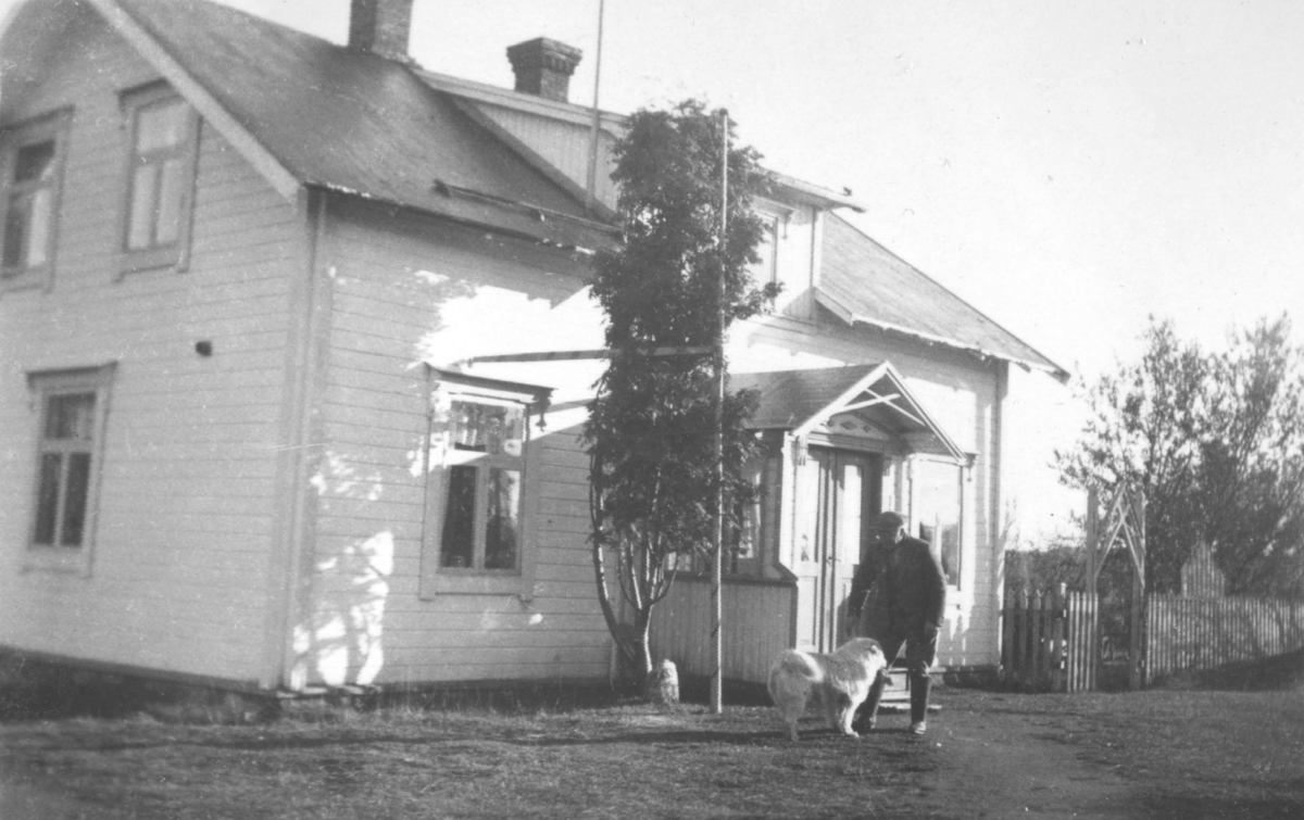 En mann med en lys hund står foran innganegen til et hus. Ved siden av inngangen et stort tre.